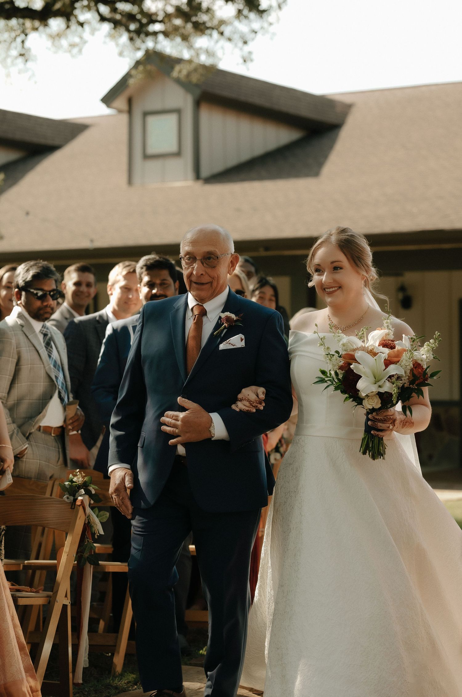 Wedding party members walk down a path outside a beige building during the ceremony.