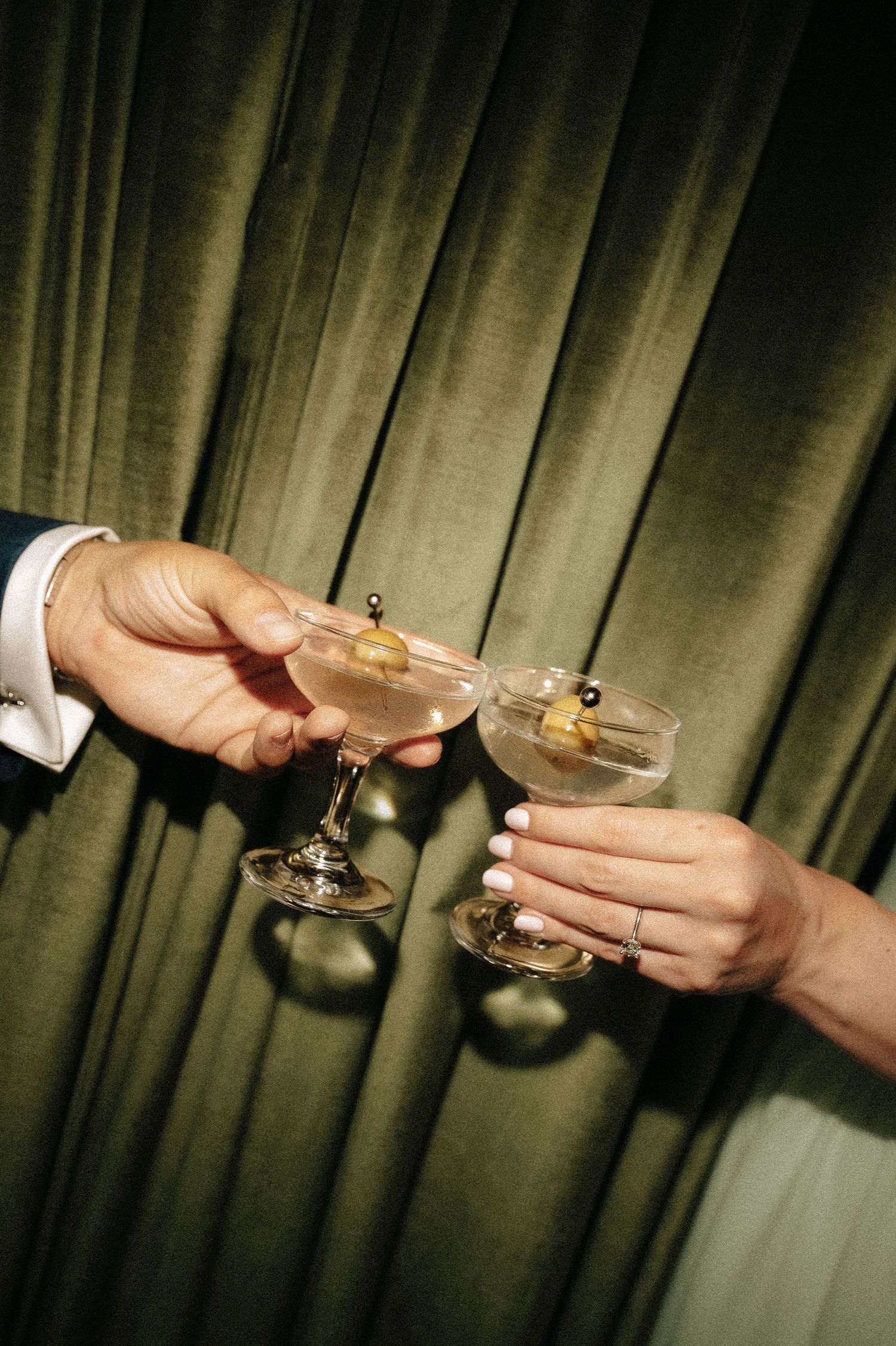 Two hands clinking martini glasses with olives against a green velvet curtain backdrop.