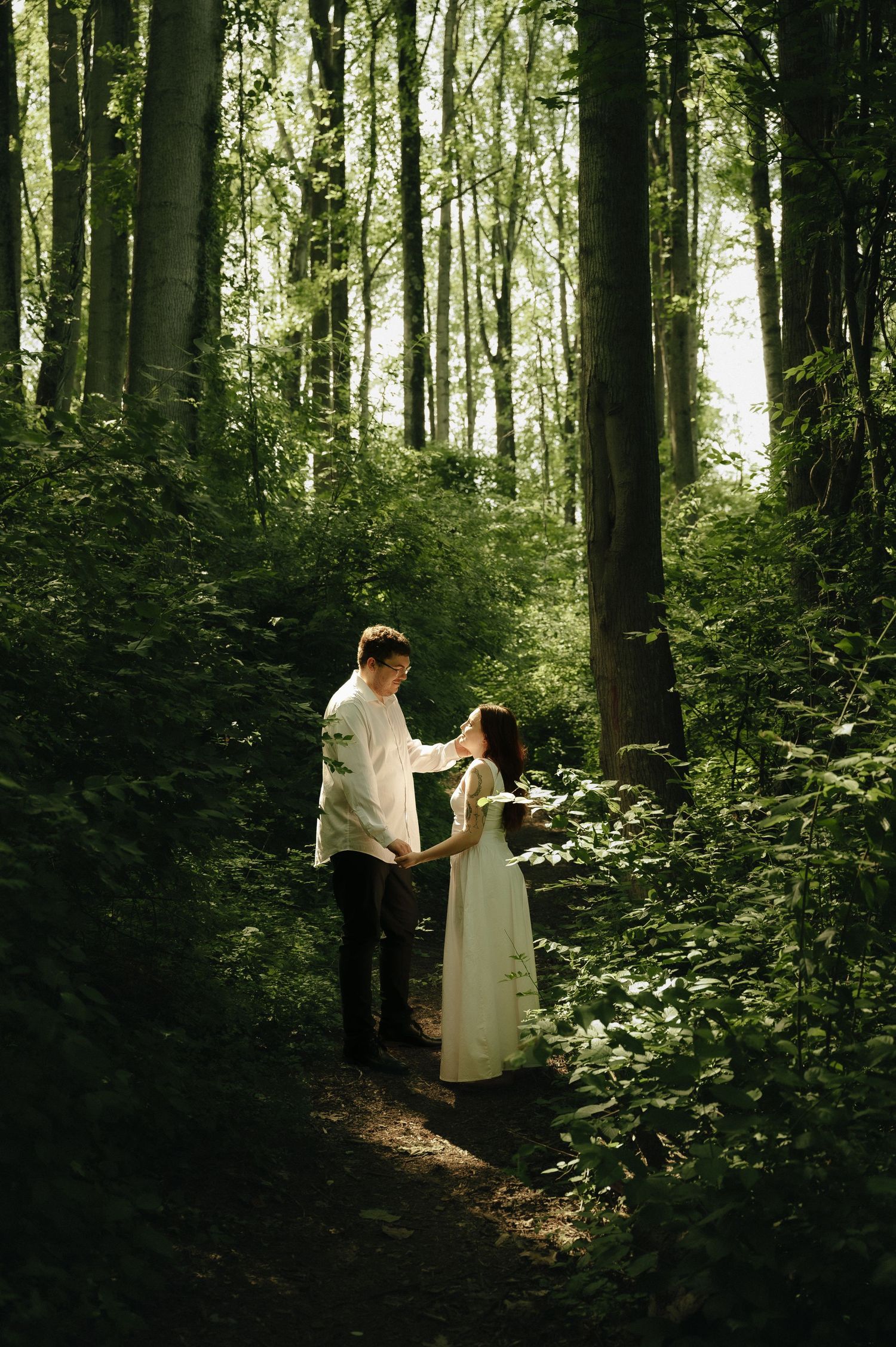 A couple in white attire holds hands on a forest path surrounded by lush green trees and sunlight.