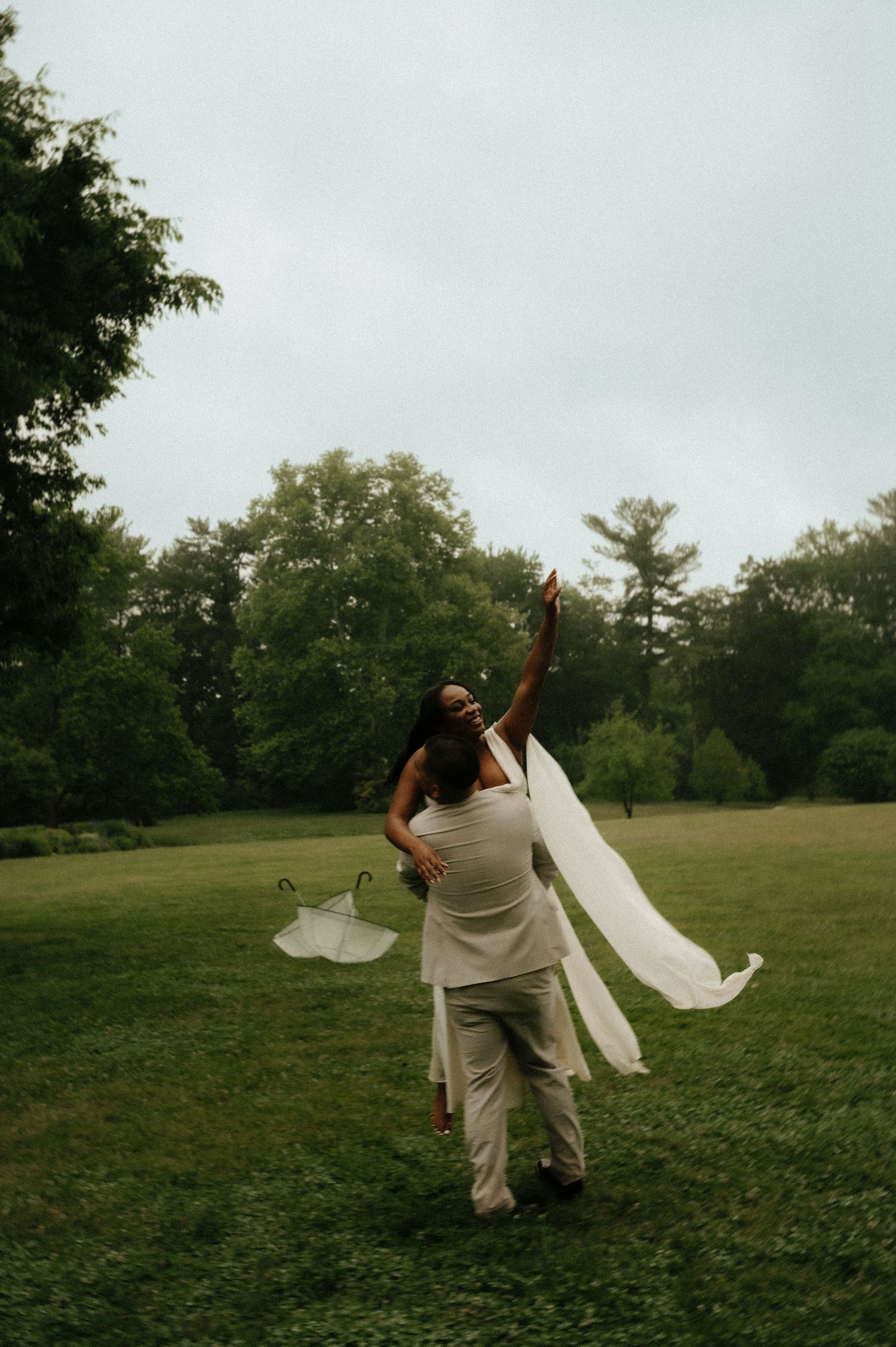 Bride and groom dancing joyfully in a lush green field, her veil flowing in the breeze on a cloudy day.