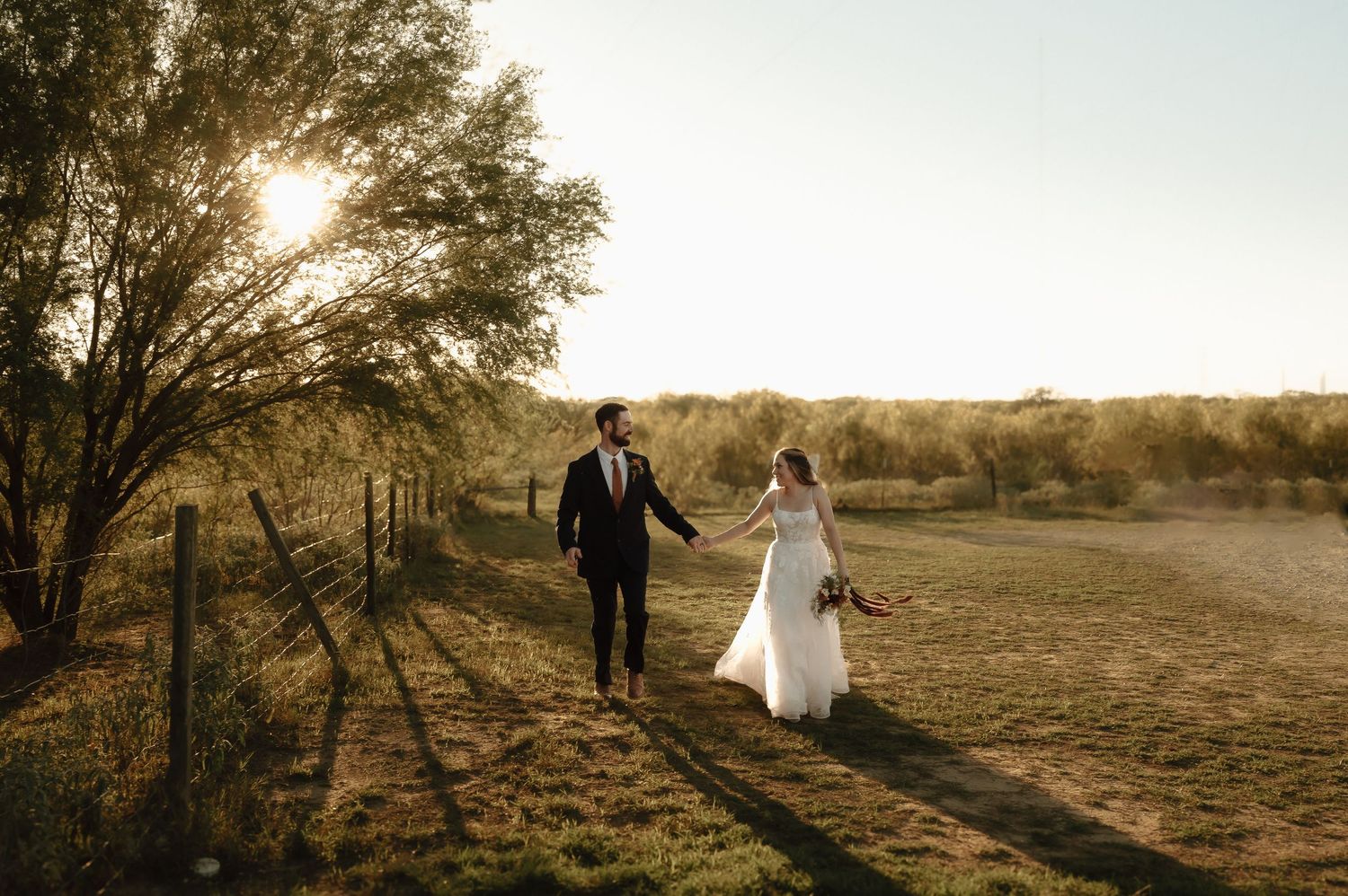 Silhouetted wedding couple walking through golden sunset field.