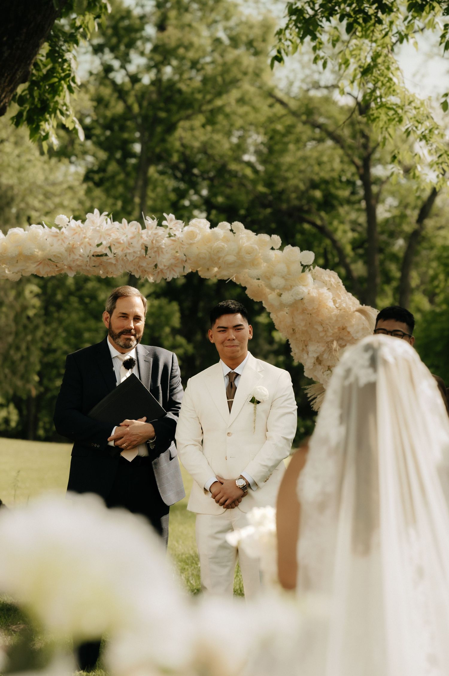 Groom in white suit smiles at bride approaching floral balloon arch at outdoor wedding ceremony.