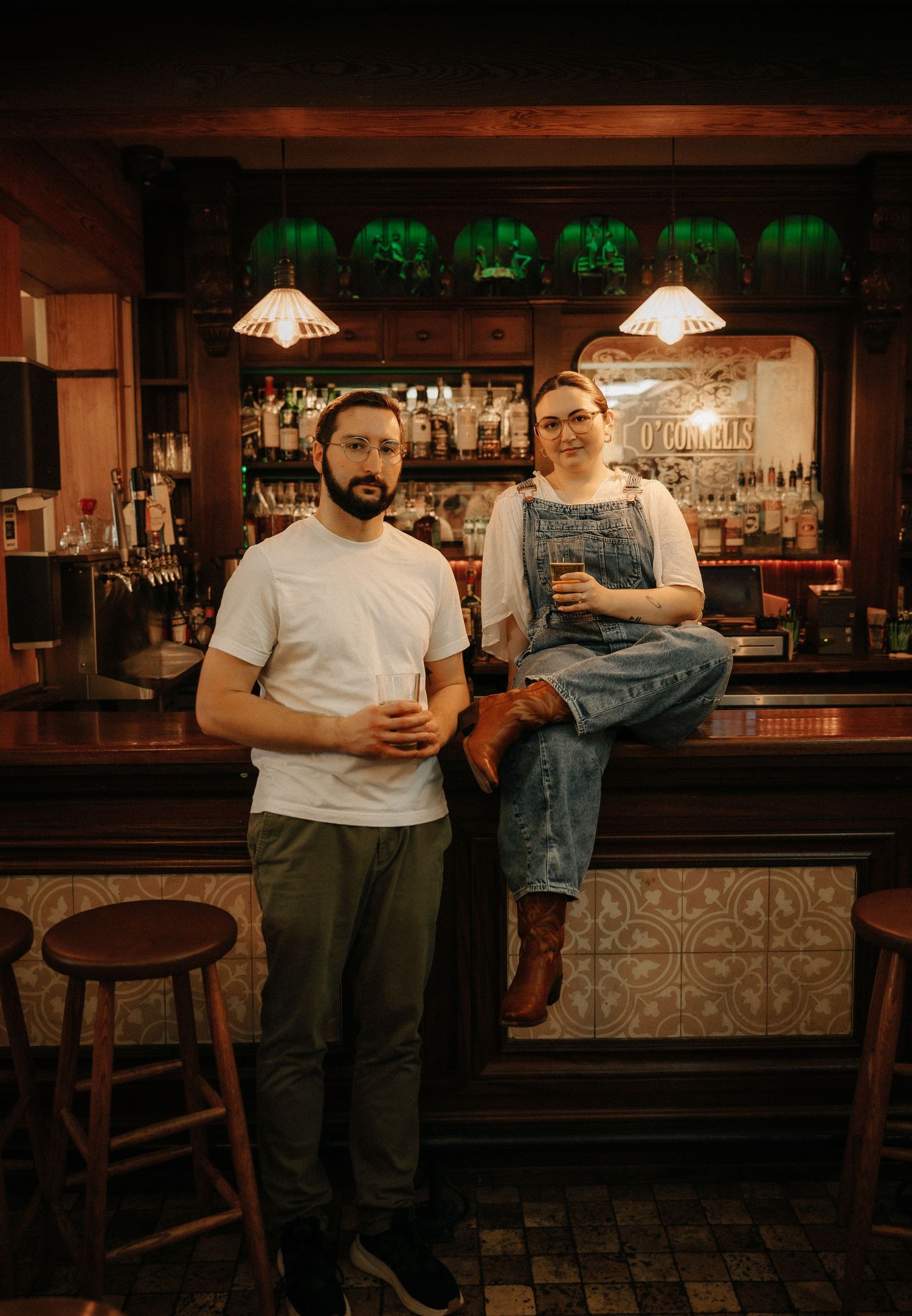 Two people smiling at an Irish pub bar, one seated on the counter in denim overalls, the other standing in a white tee.