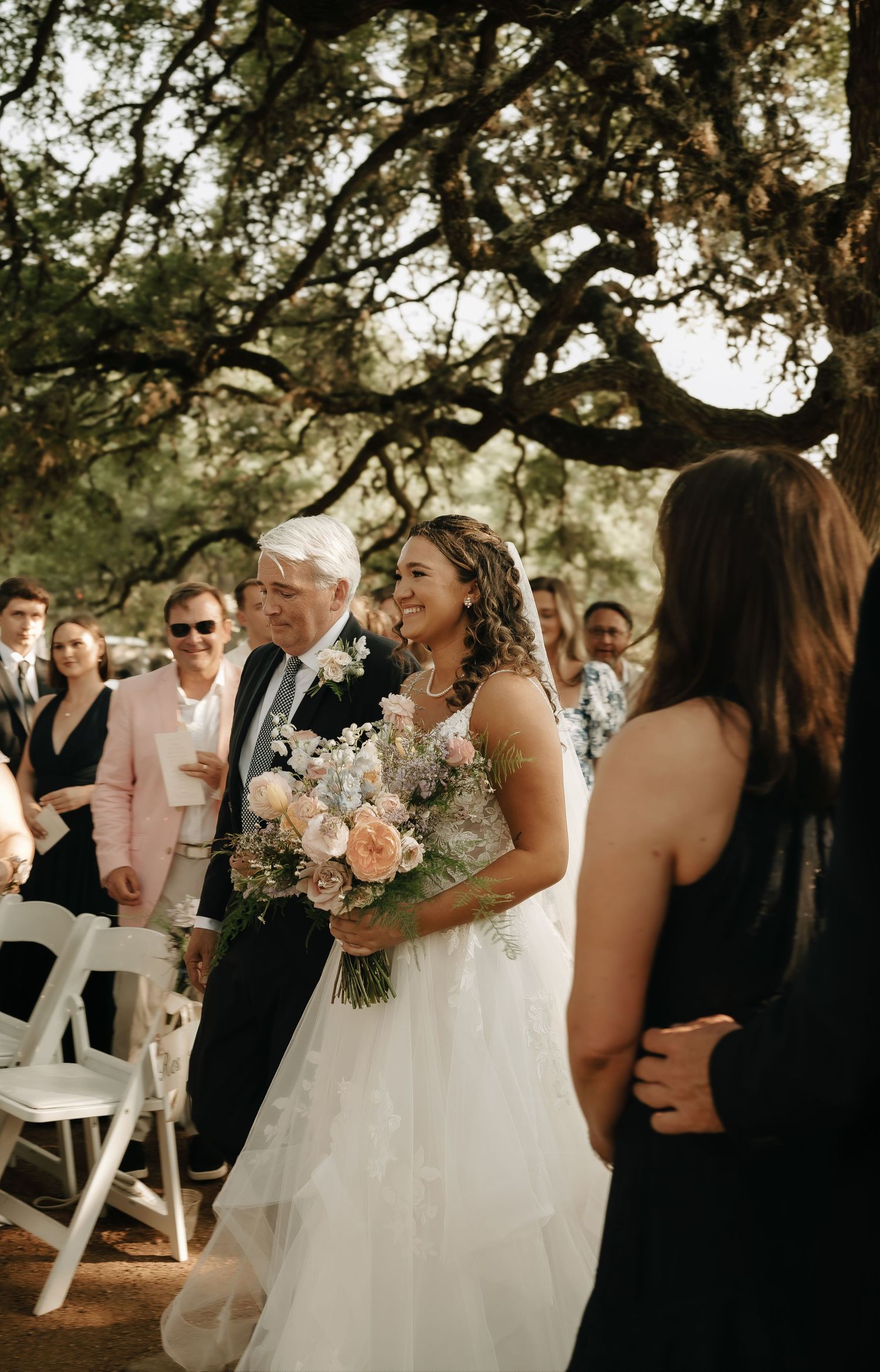Wedding guests gather under trees at an outdoor reception with natural lighting.