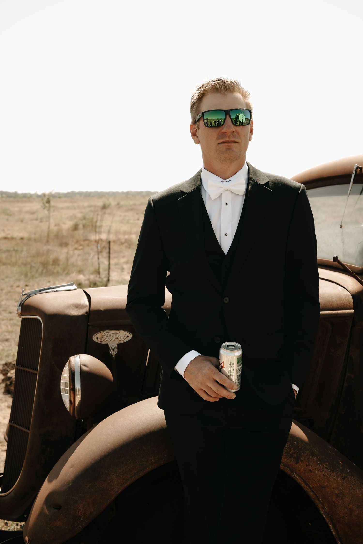 Groomsmen in black suits and sunglasses pose casually against vintage cars in a rustic outdoor setting.