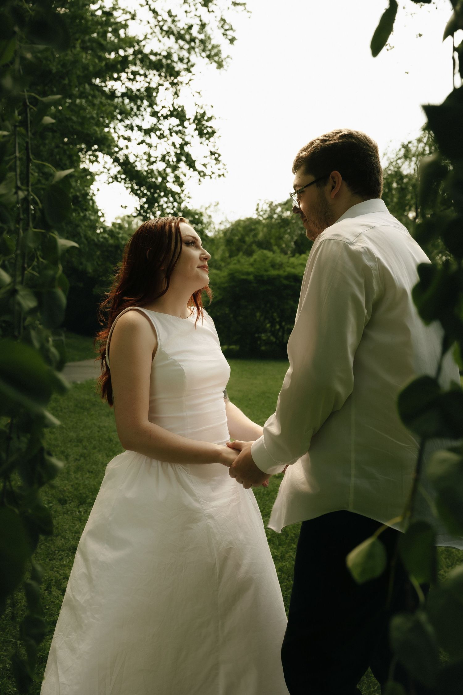 A smiling bride in a white dress holds hands with a groom in a white suit surrounded by lush green garden foliage.