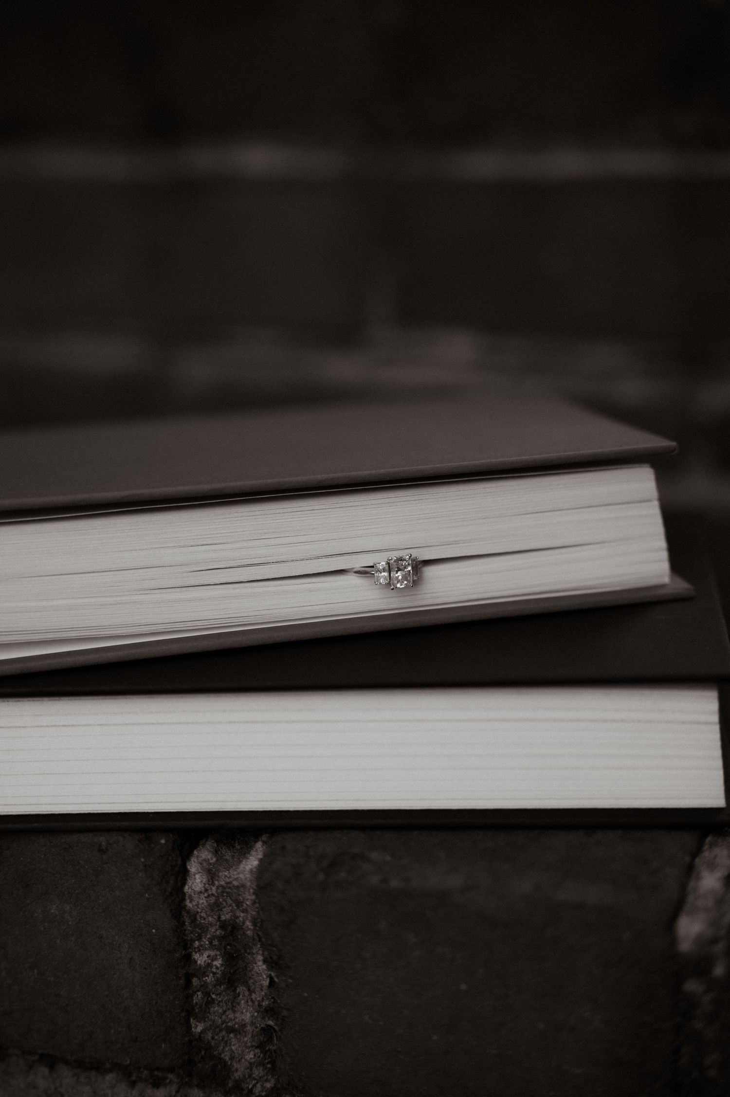 A diamond engagement ring resting between two stacked hardcover books on a dark brick surface.