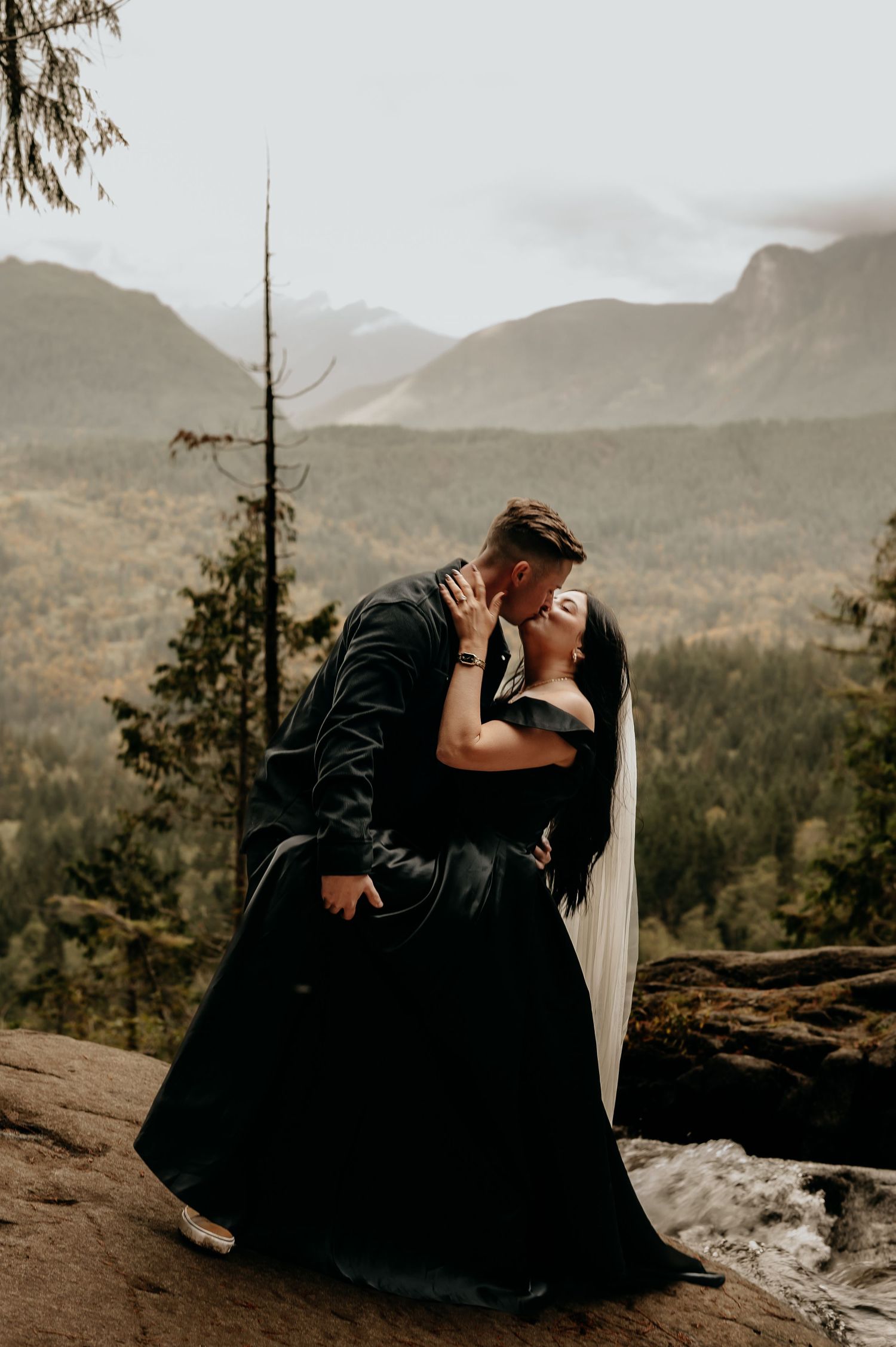 A couple shares an intimate moment on a mountain overlook with dramatic landscape views.