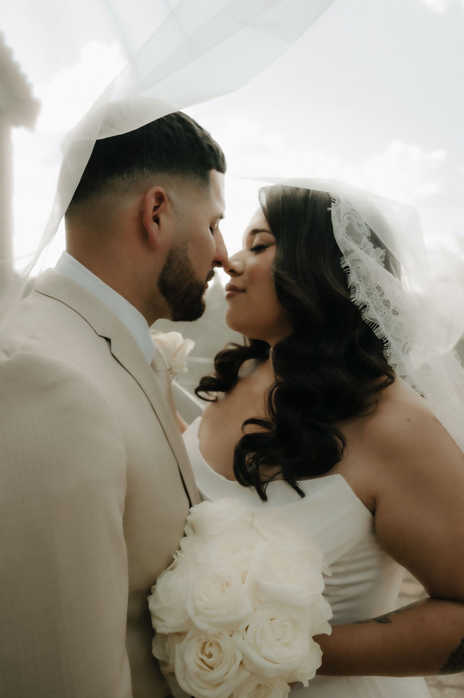A romantic wedding portrait shows a couple sharing an intimate moment with white roses and a flowing veil.
