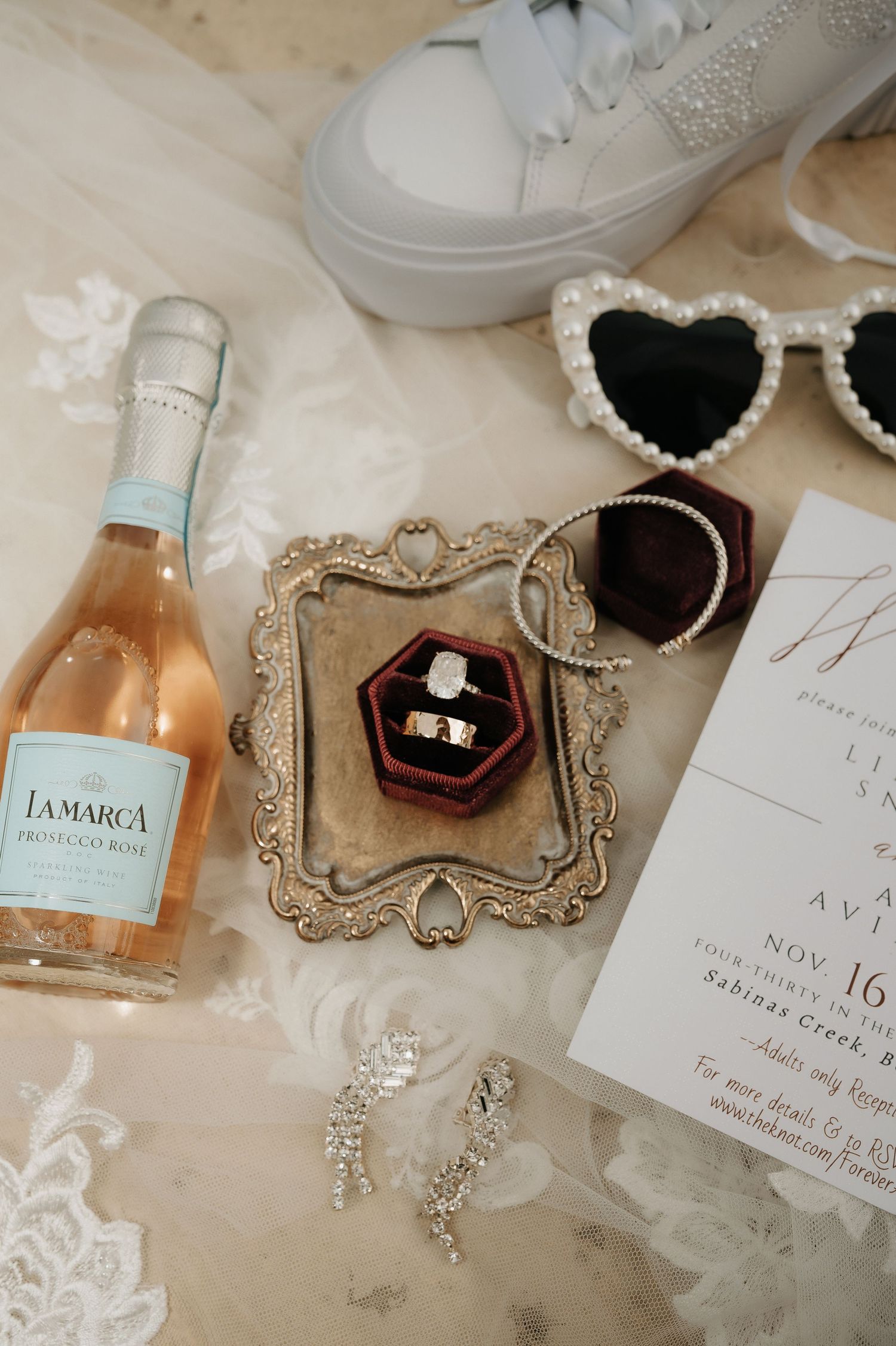 Wedding details including rings, invitations, perfume bottles, and delicate lace accents arranged on a light surface.