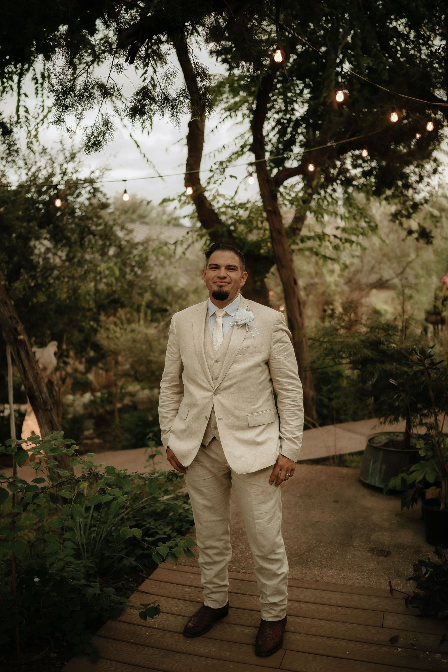 Groom in light beige suit walking along garden pathway with string lights overhead.