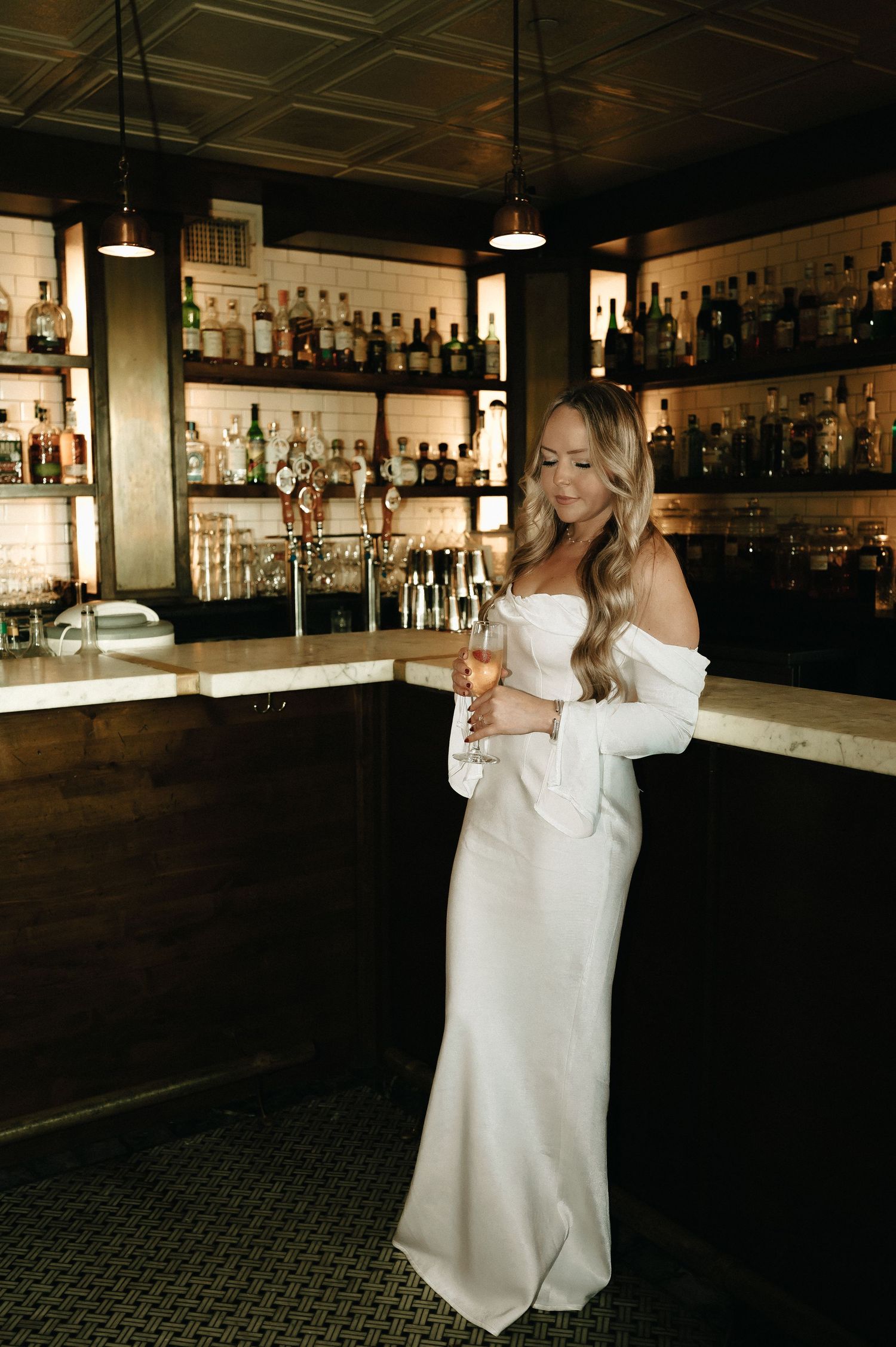 Elegant white dress against dark bar interior with backlit bottle display.