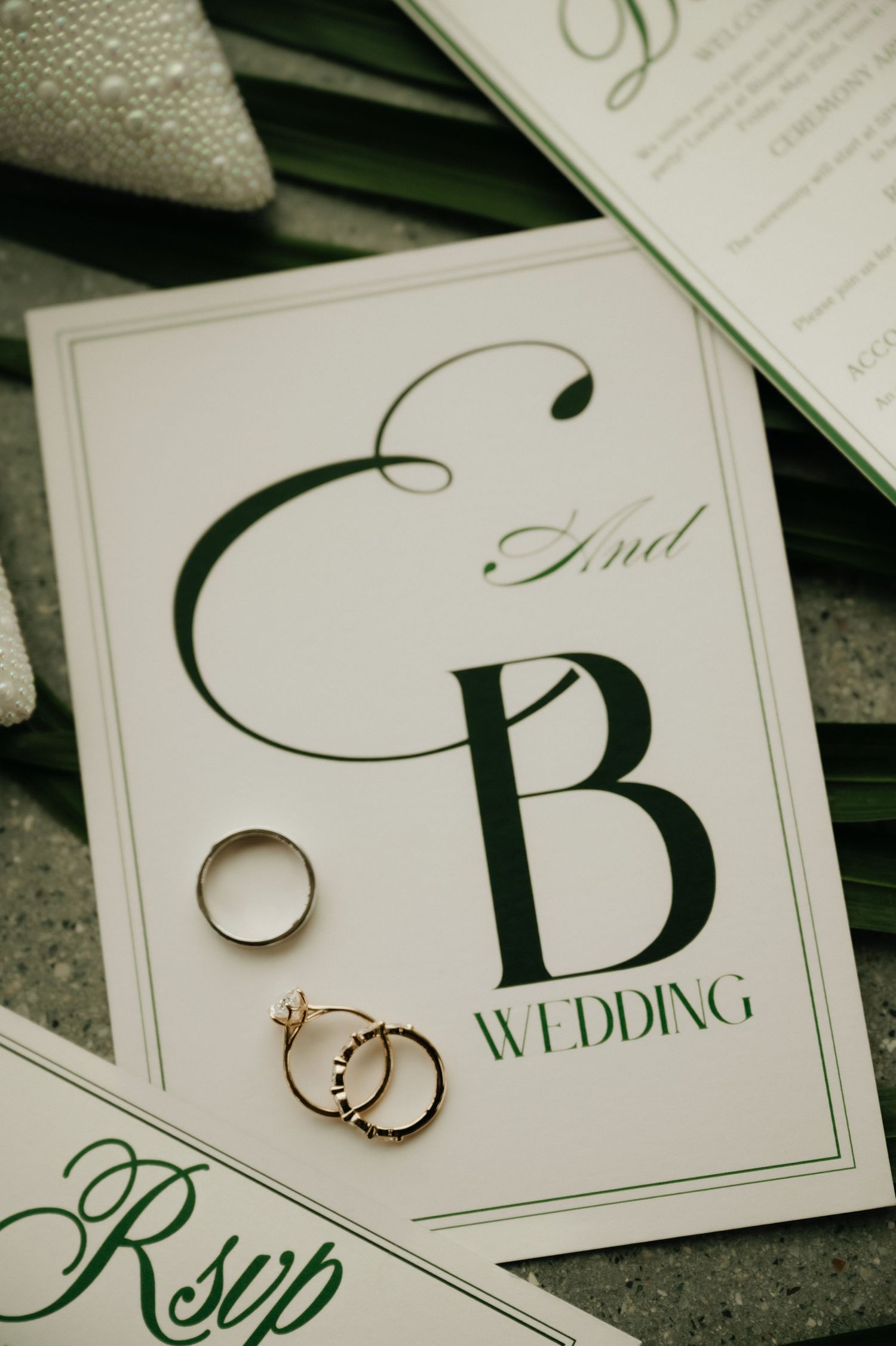 Elegant wedding invitation suite with monogram E and B, gold rings, and green typography on white cardstock.