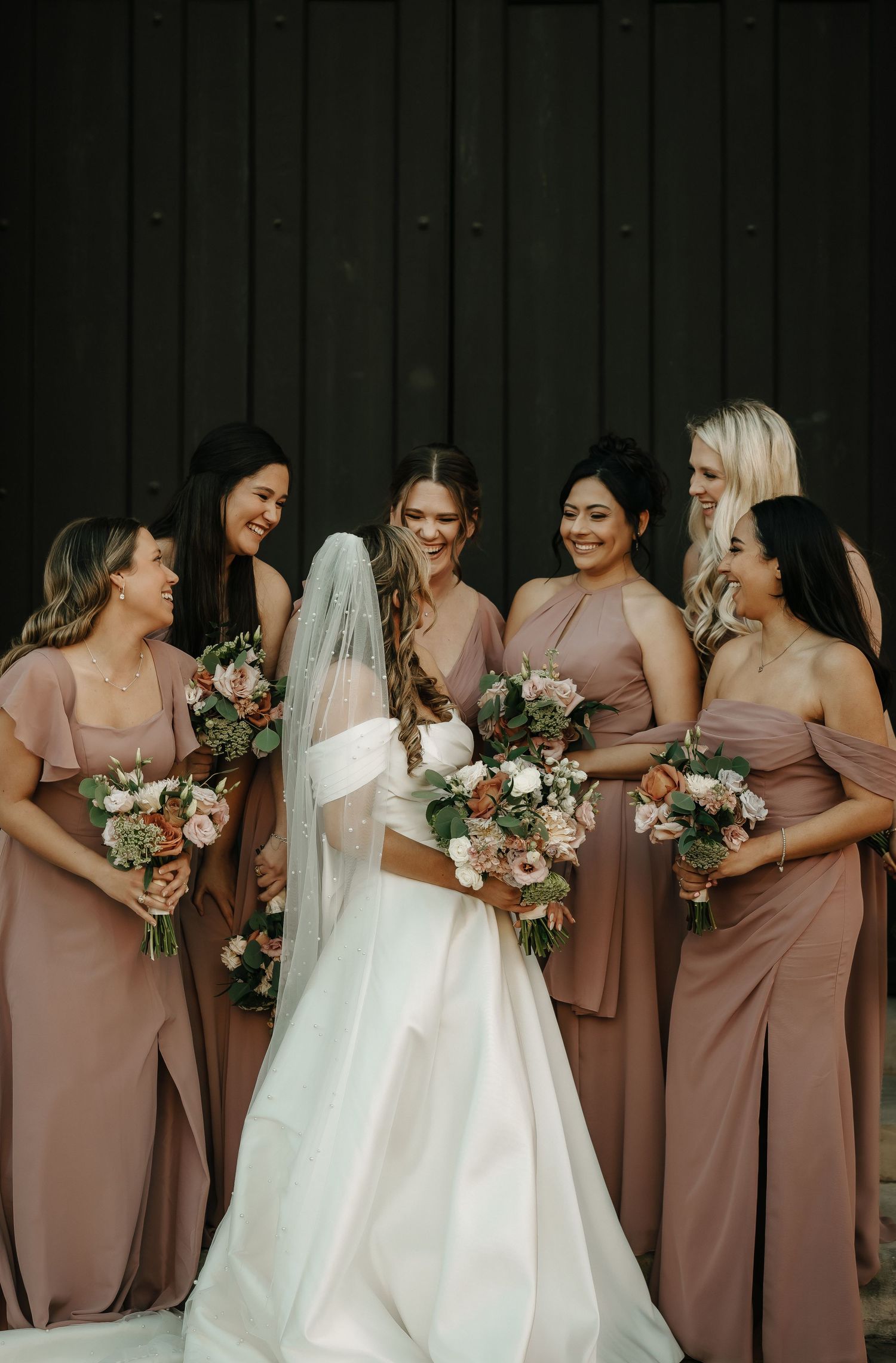 Bridal party in dusty rose dresses sharing joyful moment against dark wall background.