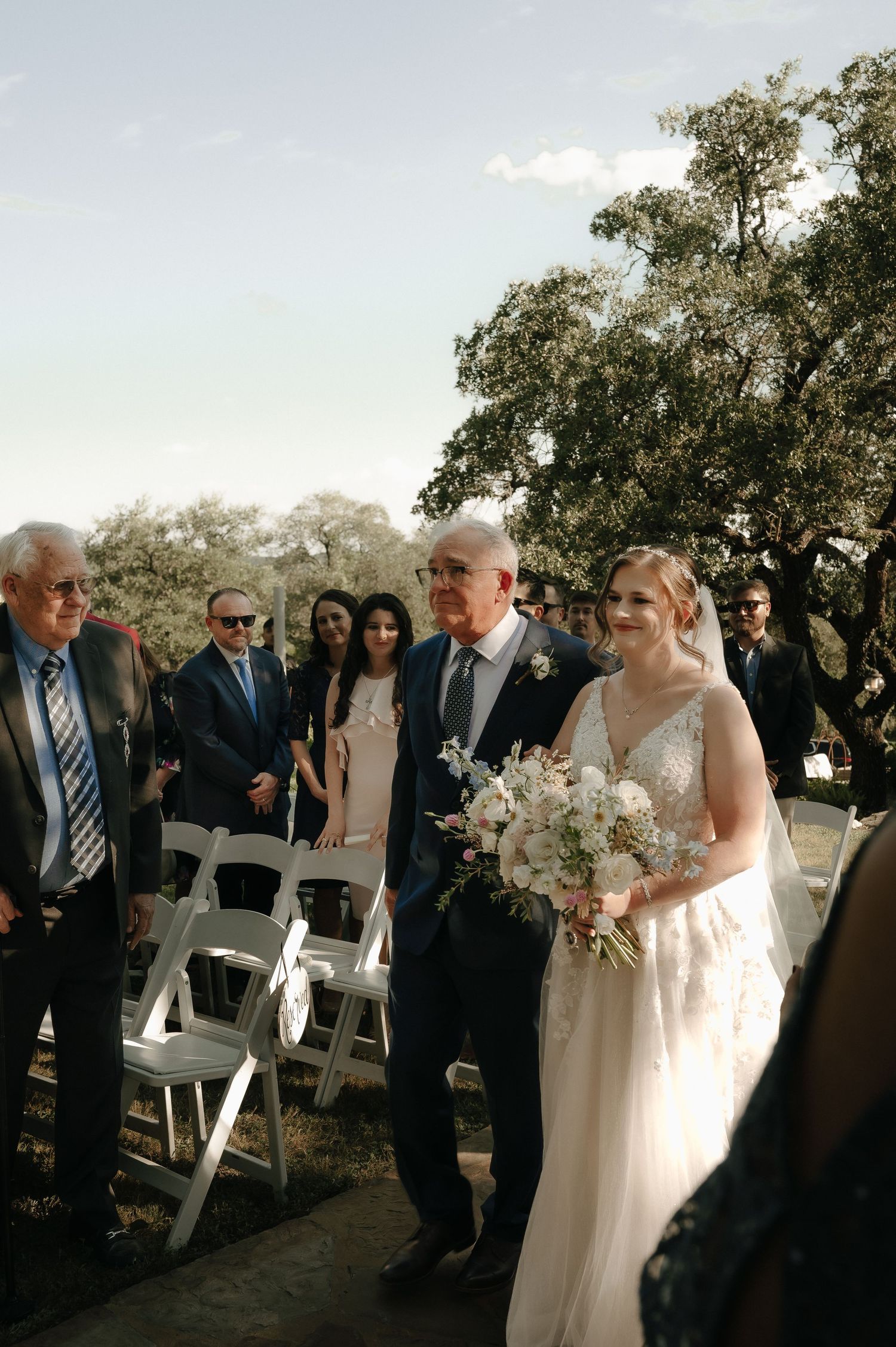 Outdoor wedding ceremony with guests seated on white chairs under tree canopy.