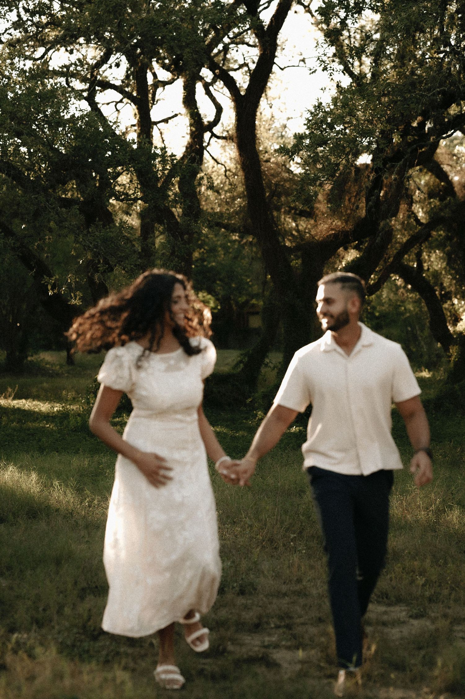 Couple walking hand in hand through a sunlit wooded area in formal attire.