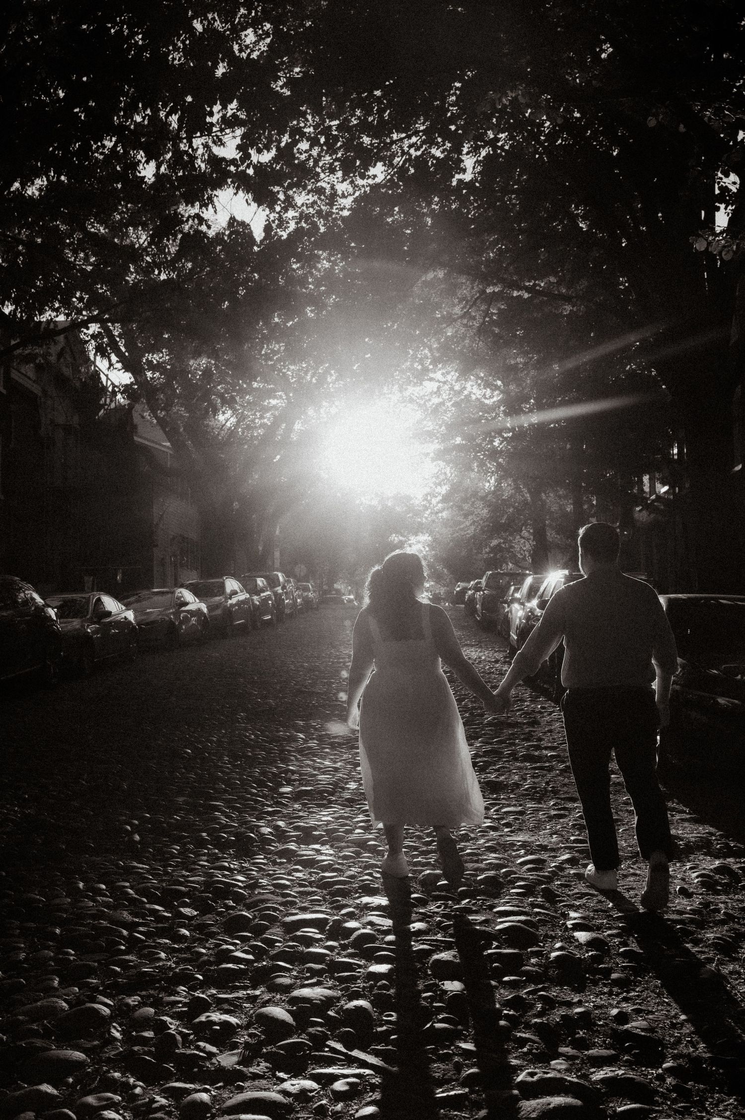 Black and white photo of a couple holding hands walking on cobblestone street toward bright sunlight through tree canopy.