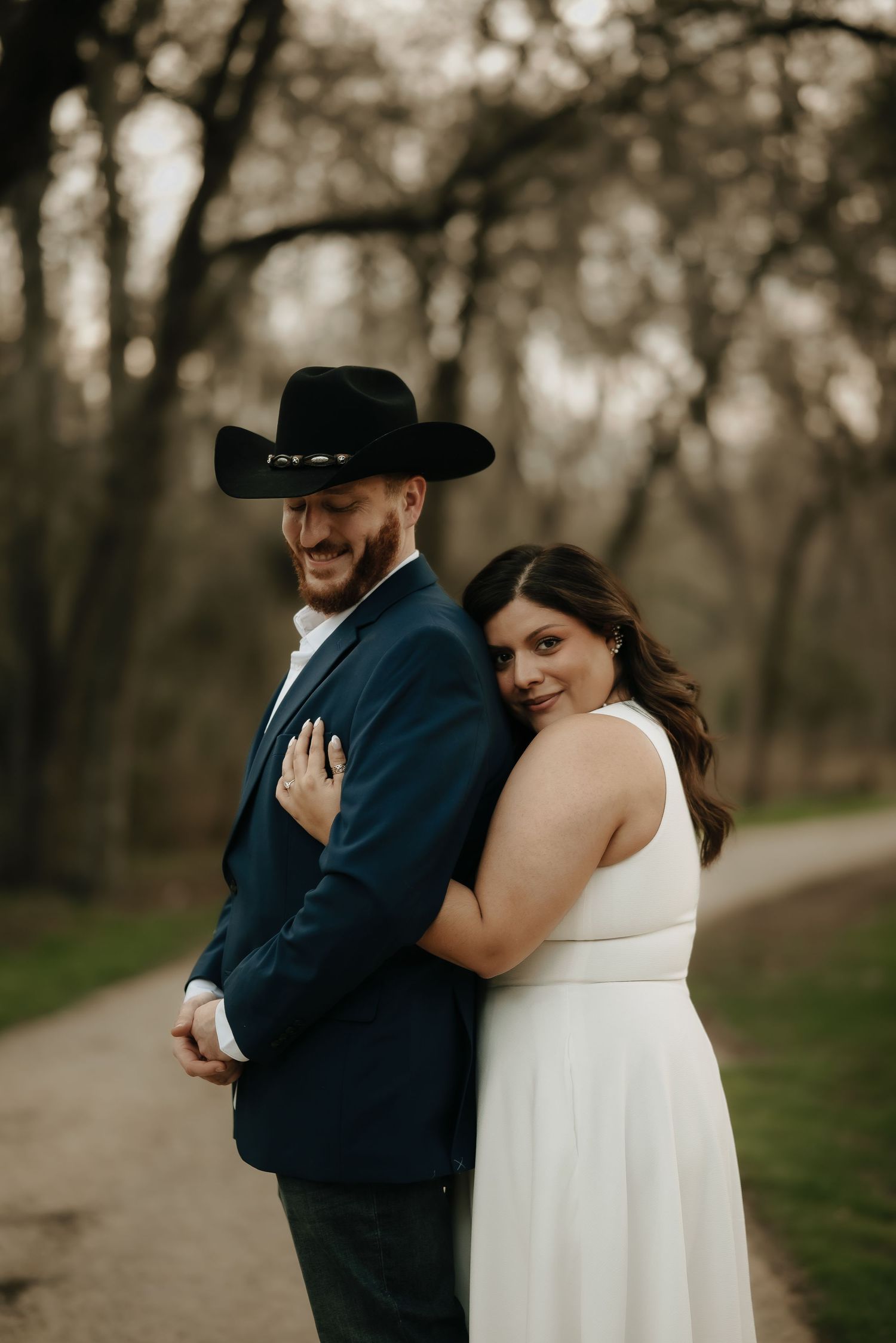 A couple in wedding attire shares a tender moment along a tree-lined path.