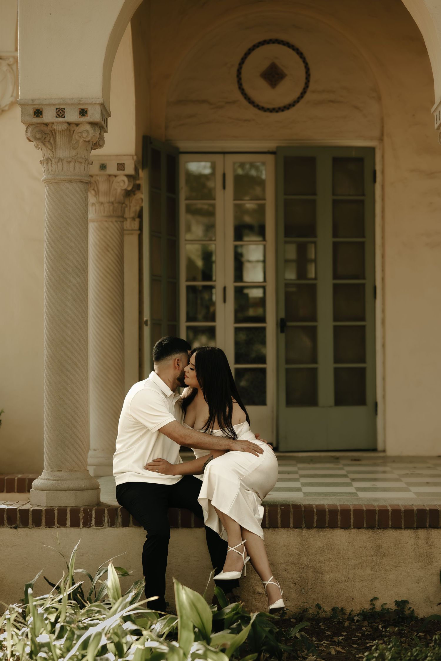 A romantic photoshoot under arched columns featuring a couple in coordinated white outfits.