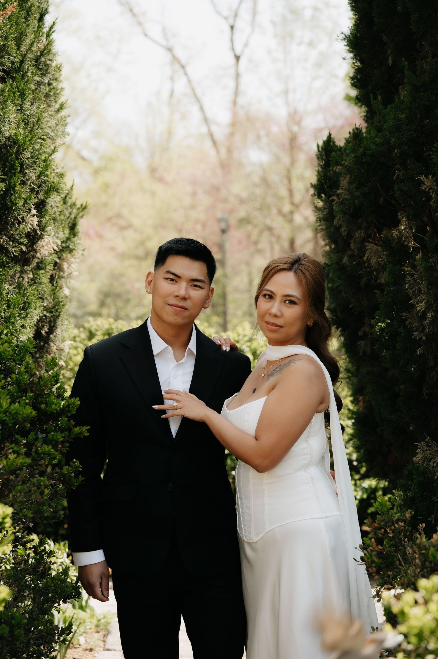Couple in wedding attire poses among cypress trees, bride in white gown, groom in black suit.
