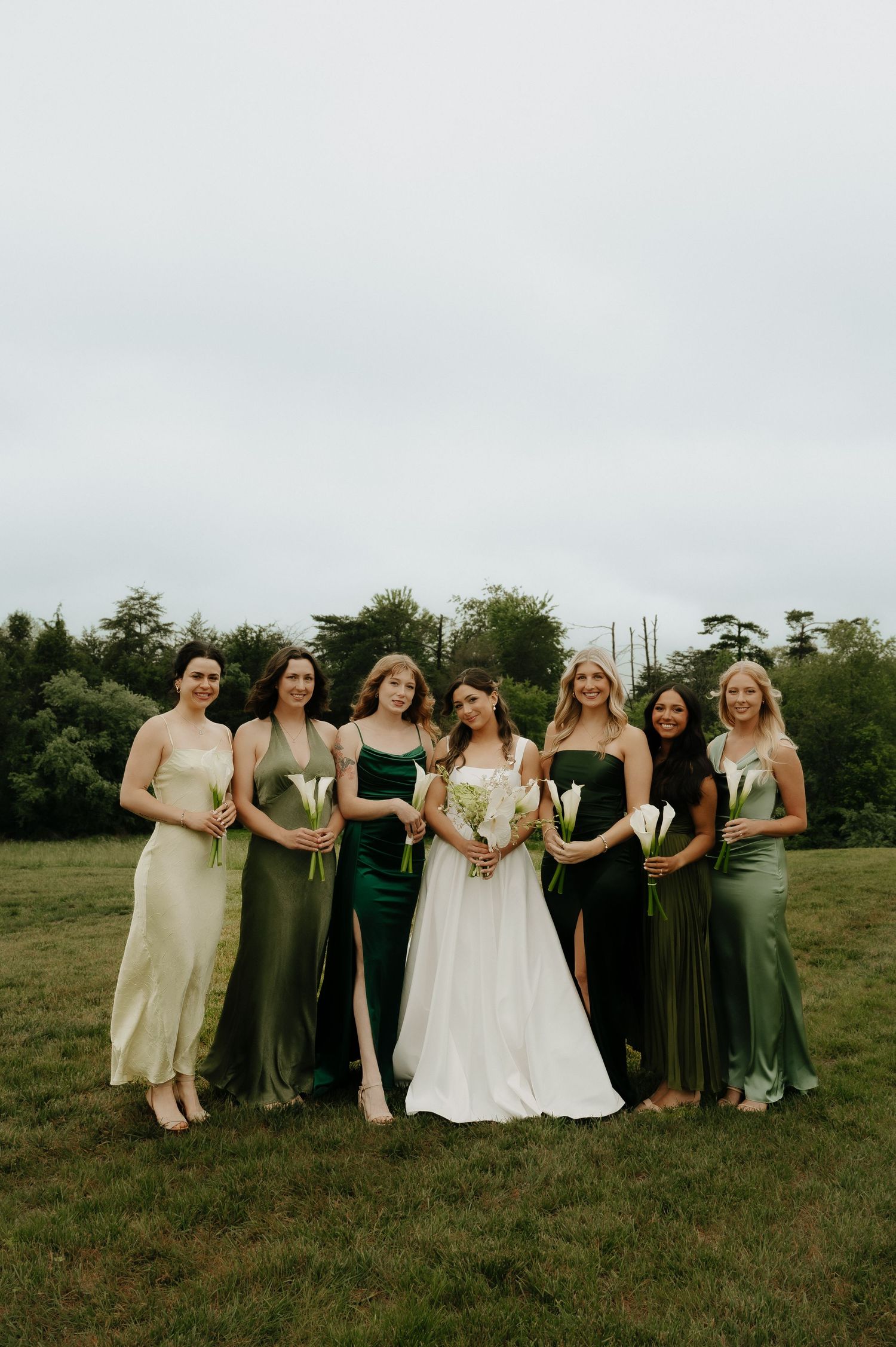 Bride in white gown poses with six bridesmaids in mismatched green satin dresses holding white calla lilies outdoors.