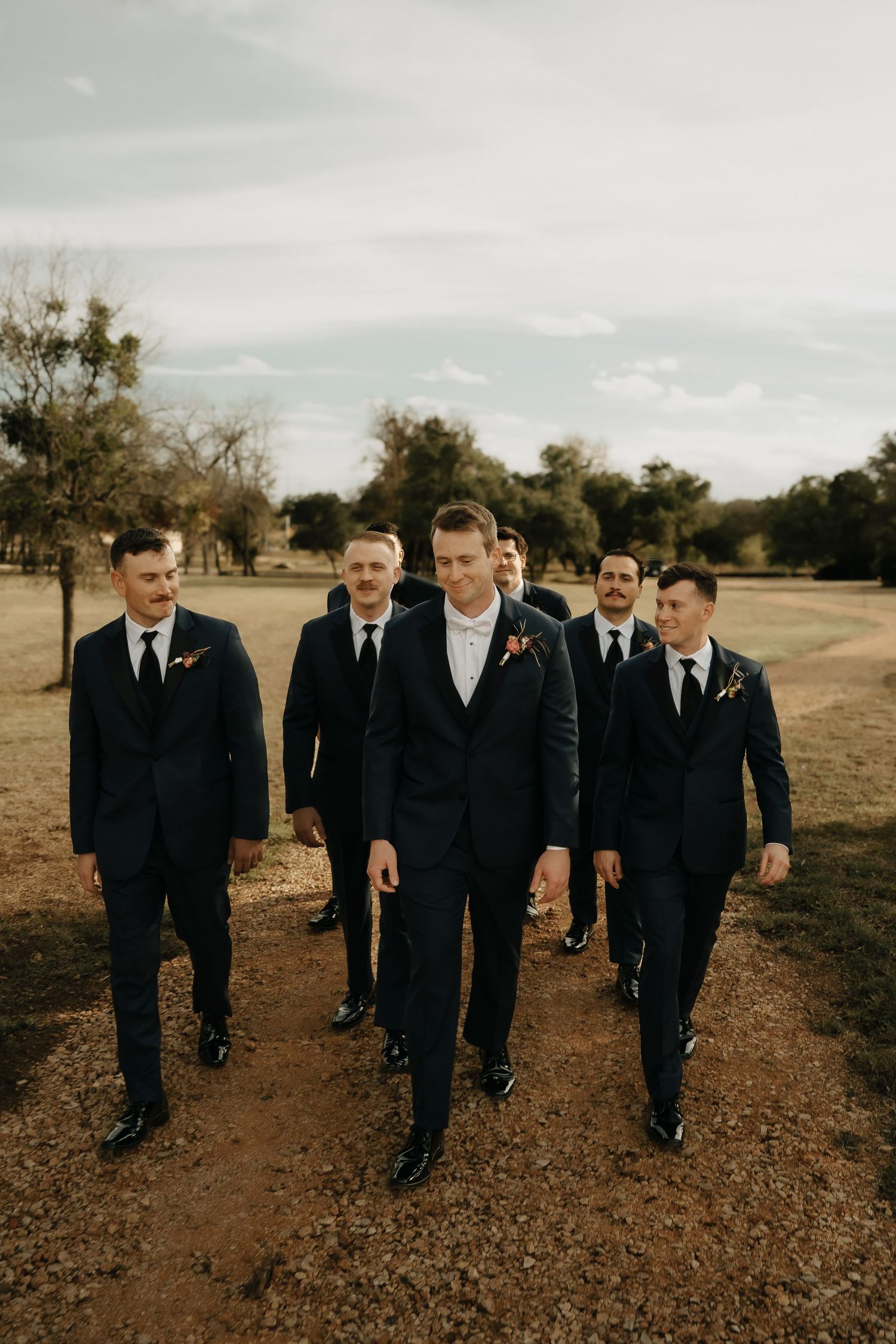 Groomsmen walk together on a dirt path wearing dark suits in an outdoor setting.