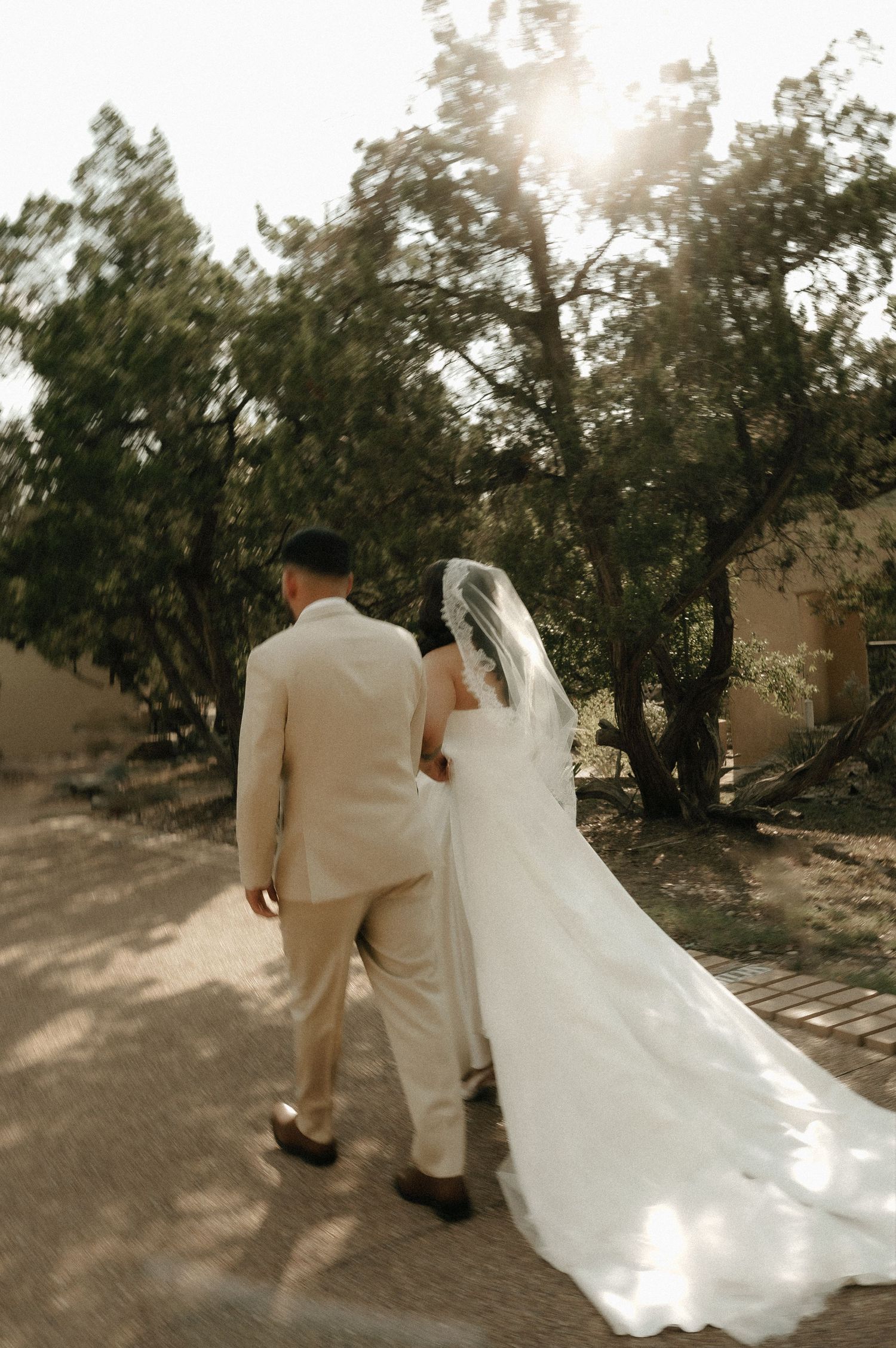 A newlywed couple walks along a garden path, with the bride's long white train trailing behind.