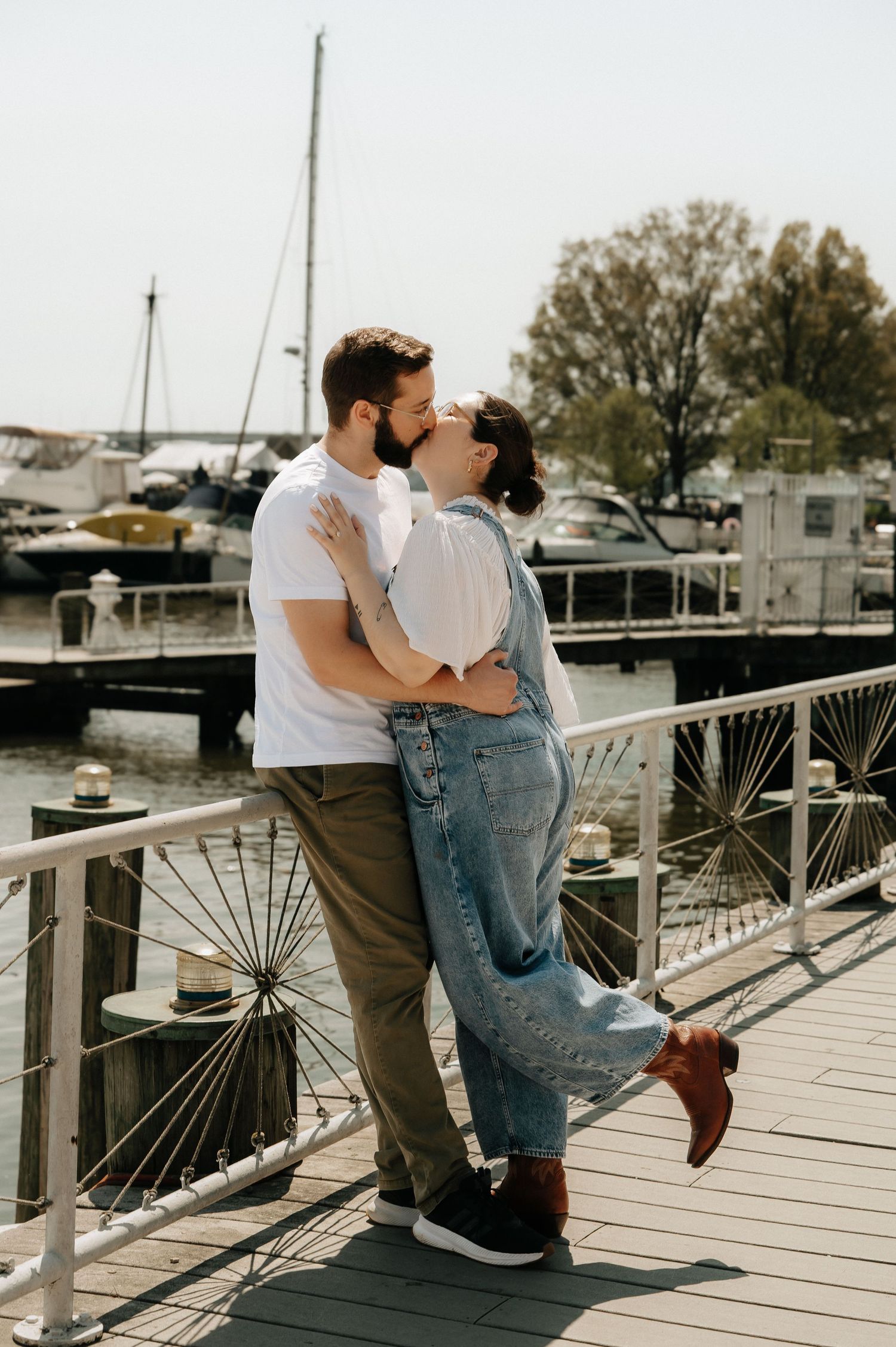 Couple sharing a romantic kiss on a marina dock, woman in denim overalls embracing man in white tee.