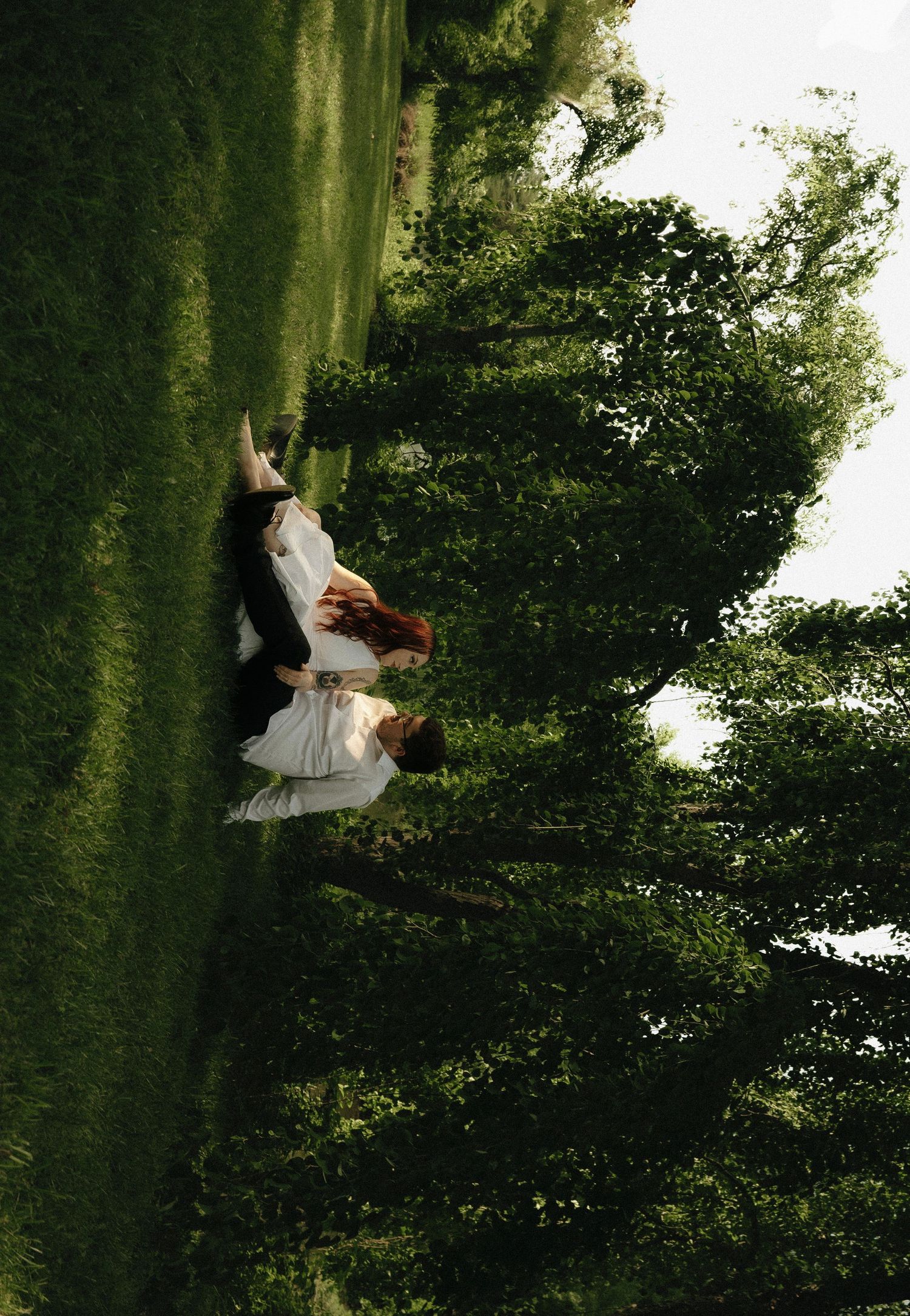 Couple embracing in a lush green park surrounded by tall trees on a bright day.