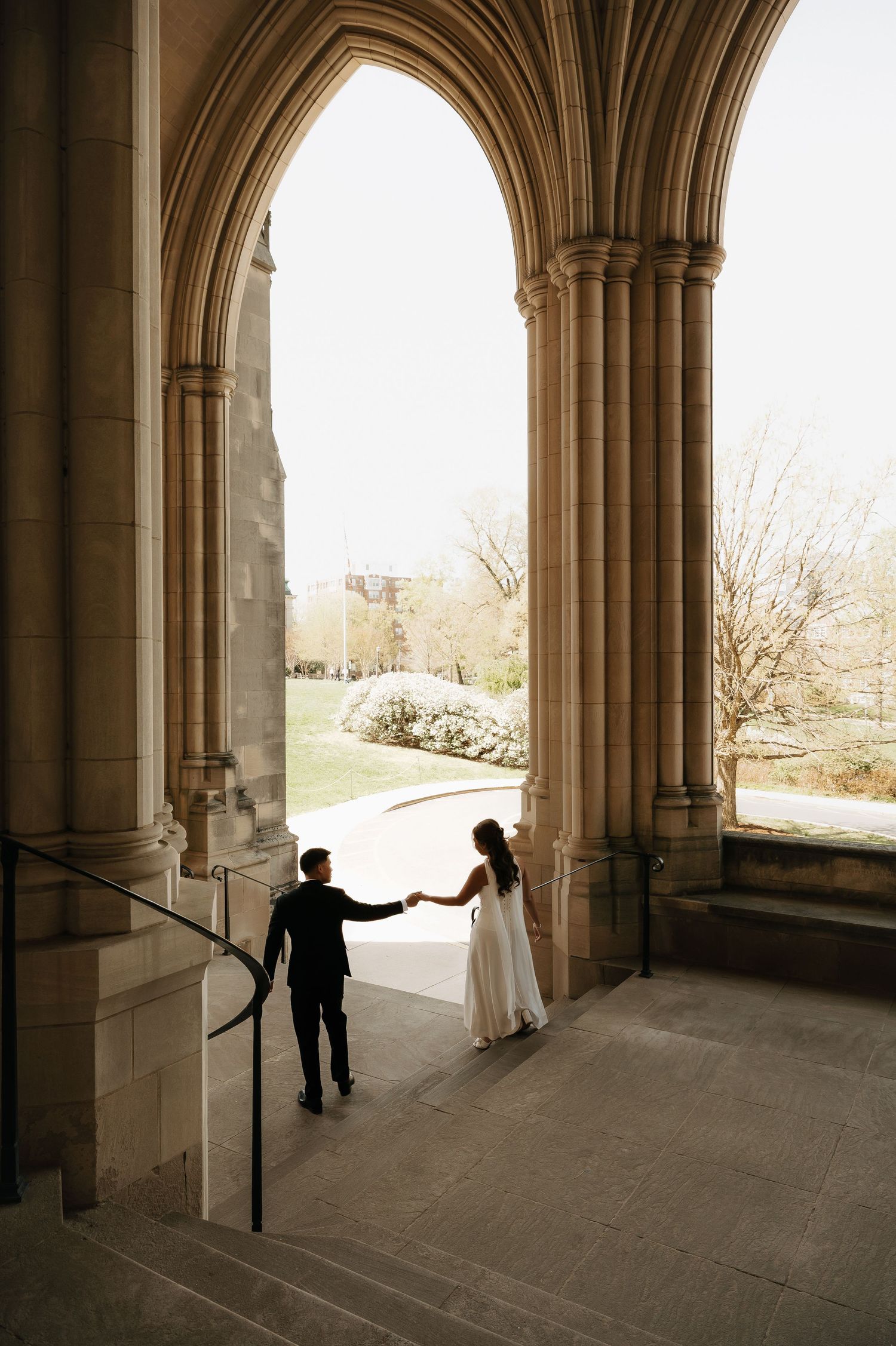 A bride and groom hold hands under a grand stone Gothic arch, walking down steps toward a sunlit garden.