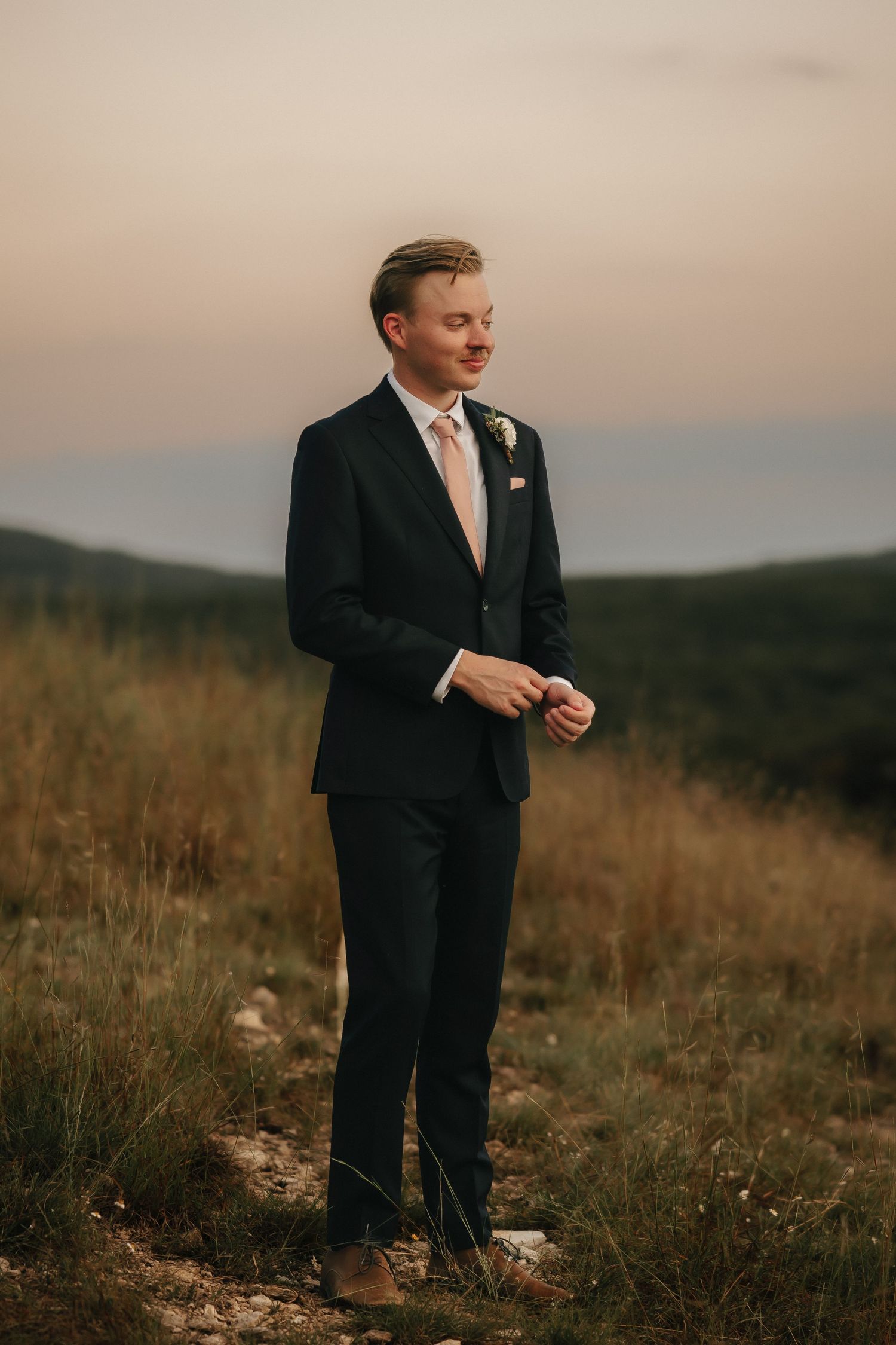 Groom in classic black suit and blush tie stands on hillside during golden hour sunset.
