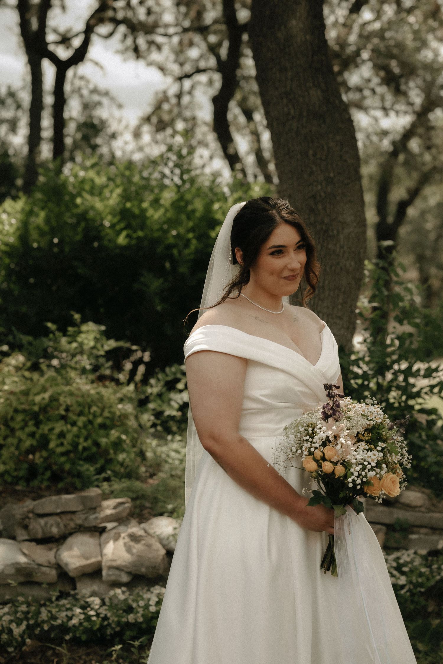 A bride holds a beautiful bouquet of mixed flowers in a natural outdoor setting.