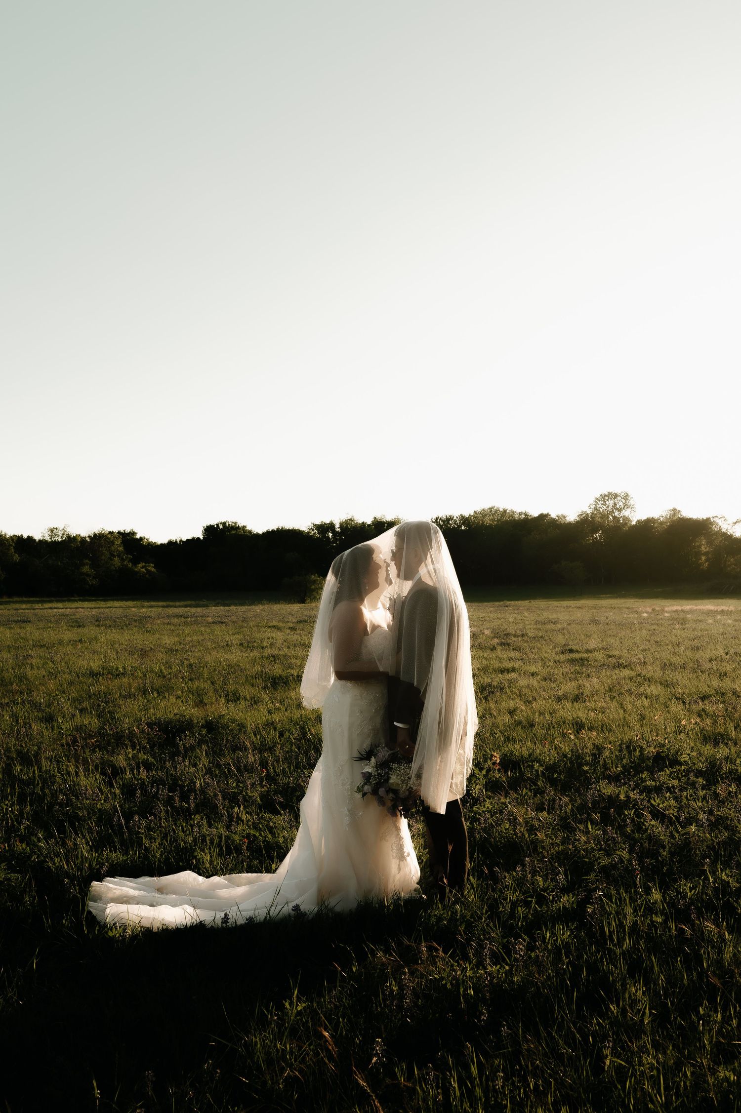 Silhouetted wedding couple in a sunset field with flowing white dress and veil.