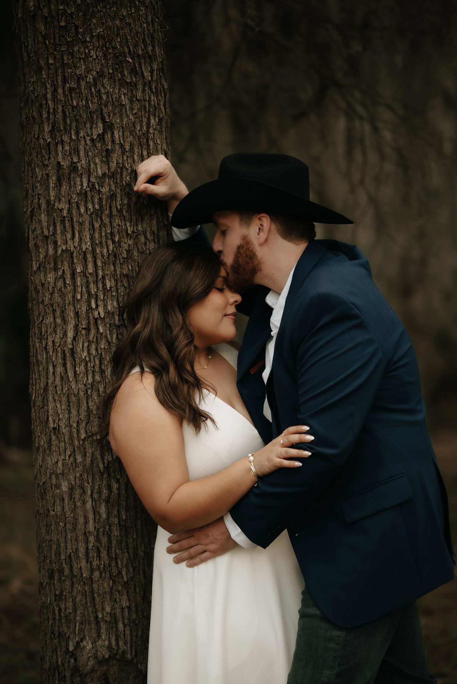 A couple in formal wear embraces against a large tree trunk in a moody forest setting.