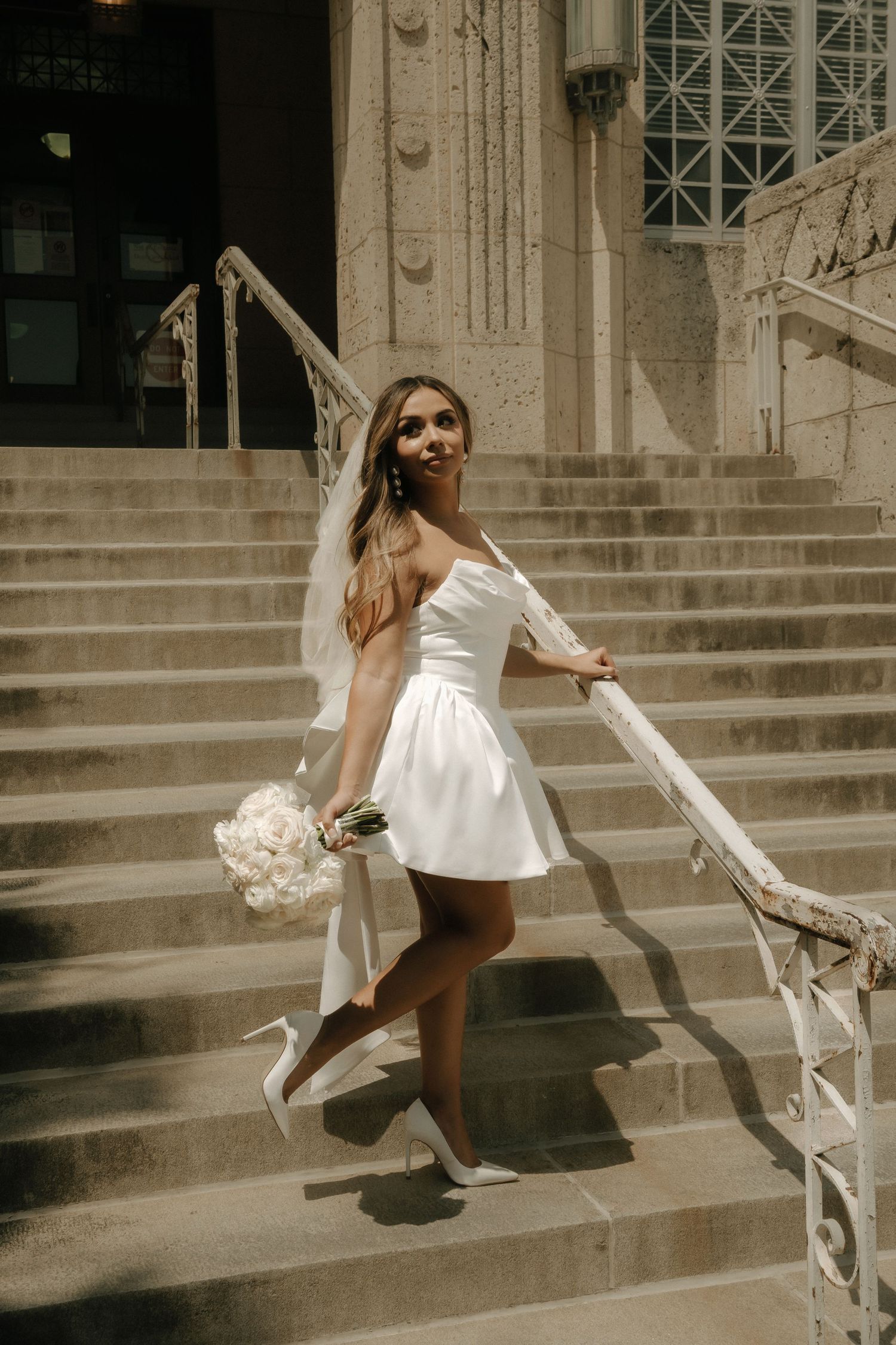 A bride in a short white dress poses elegantly on stone steps with a white bouquet.