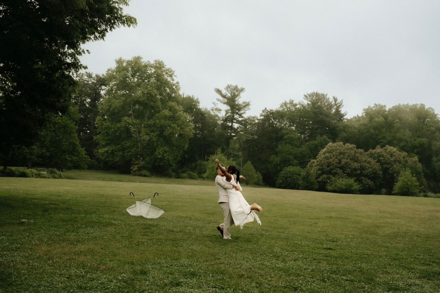 Couple dances joyfully in green meadow, bride in white gown with veil flowing, groom in tan suit.
