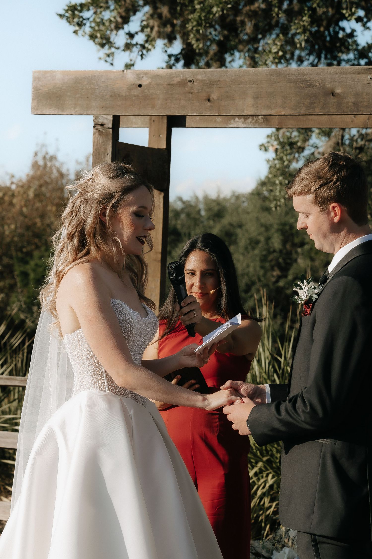 Wedding couple exchanges rings during ceremony under rustic wooden structure.