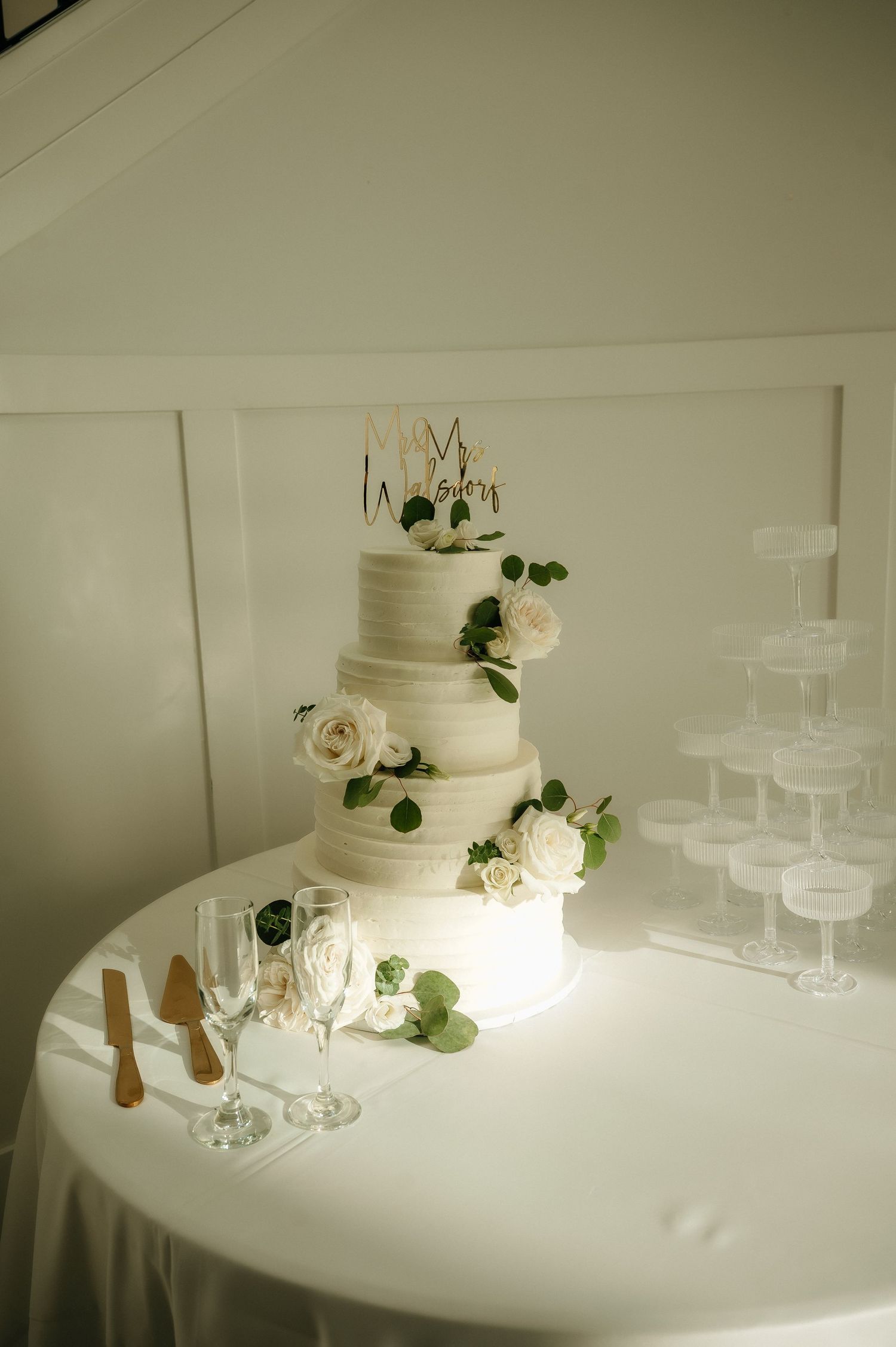Elegant white wedding cake with champagne tower display on white tablecloth.
