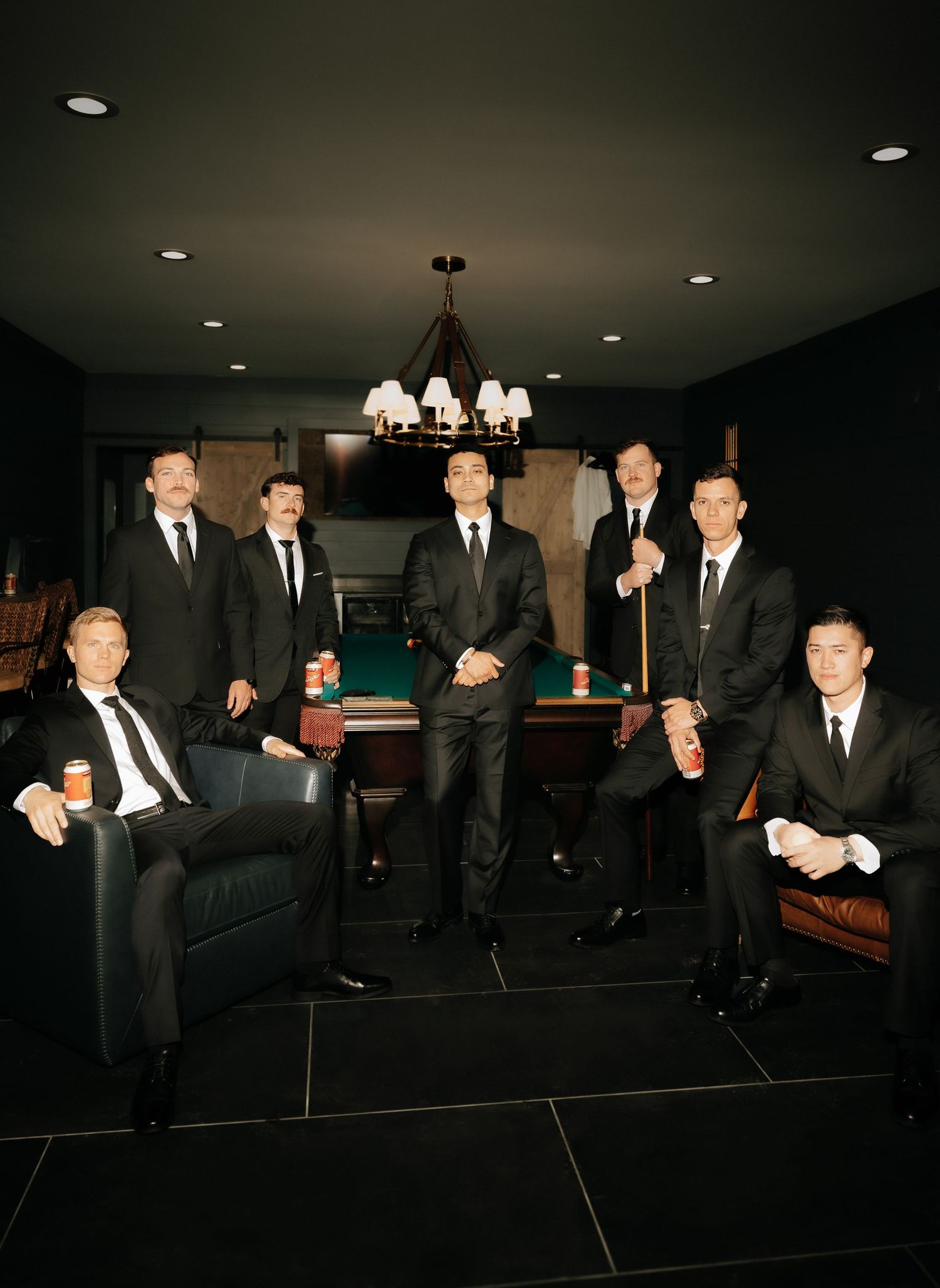 Seven men in black suits pose elegantly in a dark billiards room with a chandelier and pool table.