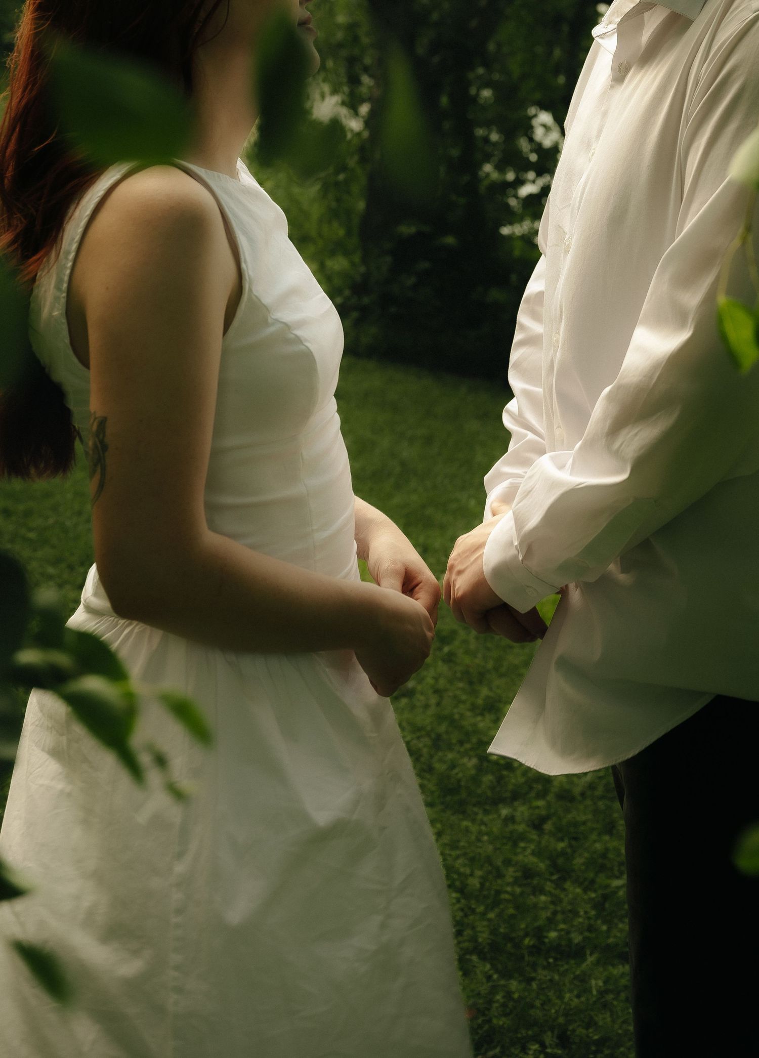 Couple in white attire holding hands outdoors in a lush green garden setting, possibly at a wedding ceremony.