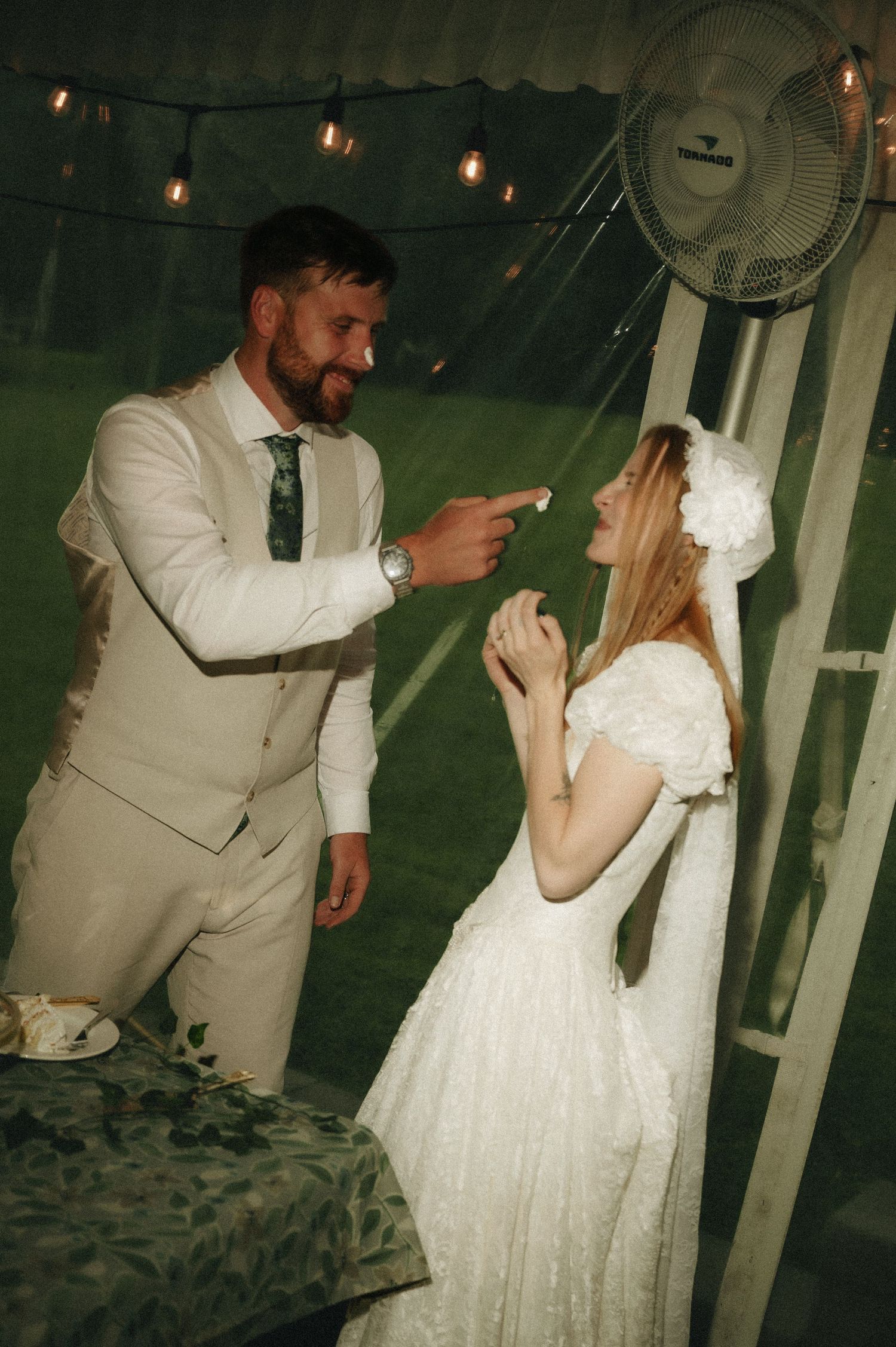Bride in white lace dress and groom in beige suit share a playful moment under festoon lights in a tent.