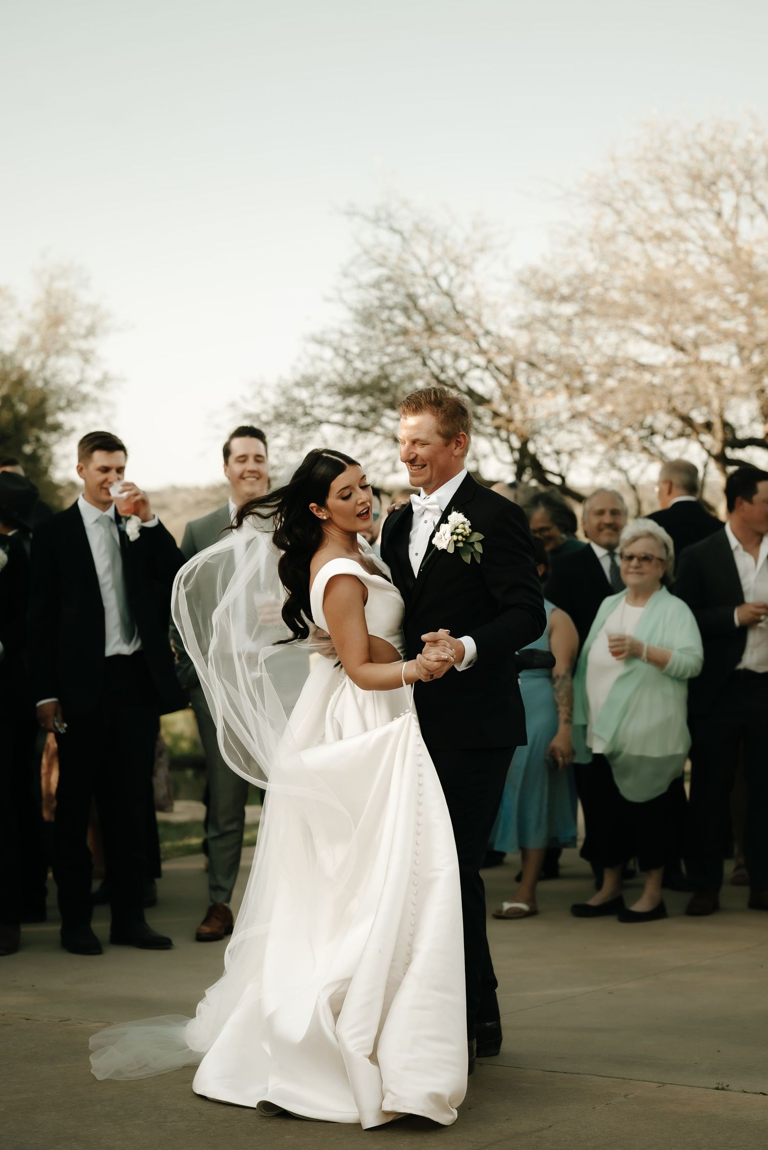 Wedding couple dancing together outdoors surrounded by guests at sunset.