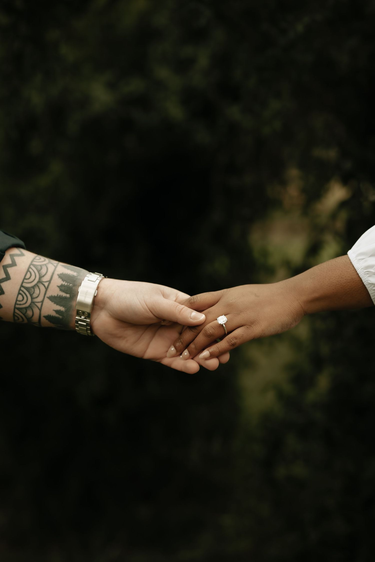 Two hands with tattoos reach out to hold each other against a dark background.