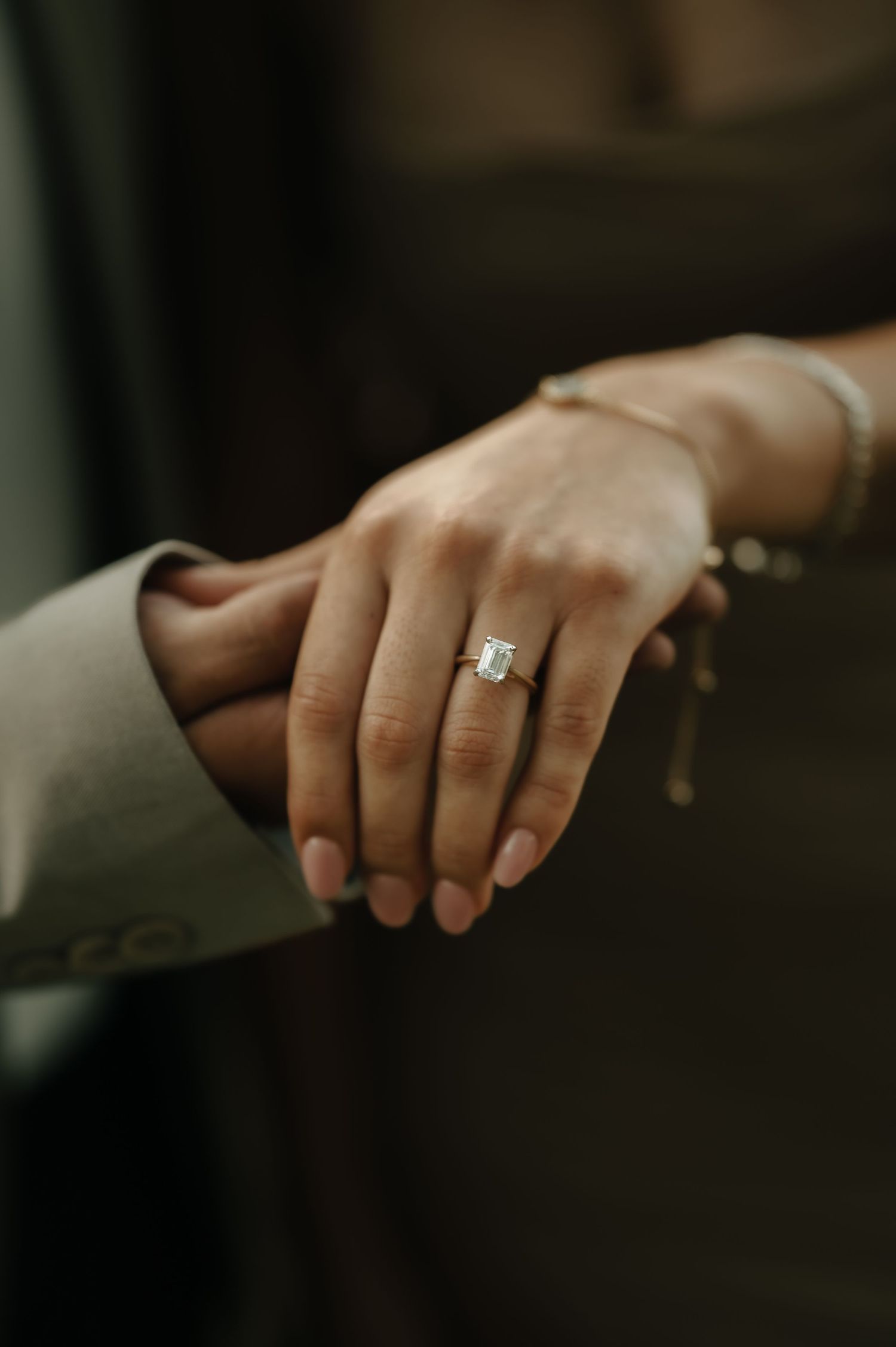 Close-up of hands with engagement ring resting on grey fabric in moody lighting.