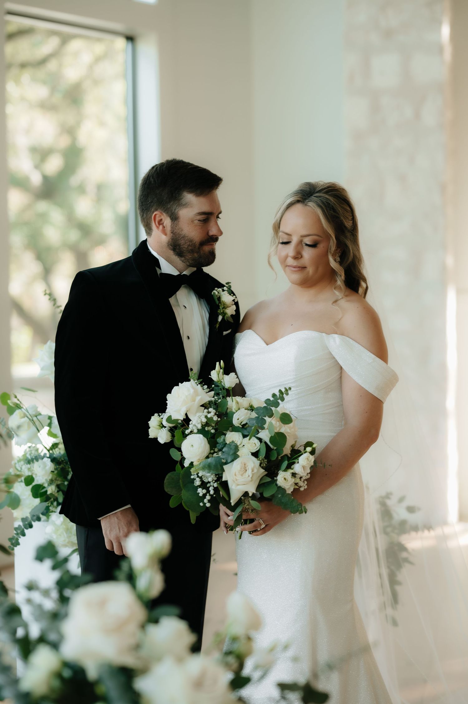 Elegant wedding couple posing with white floral bouquet near bright windows.