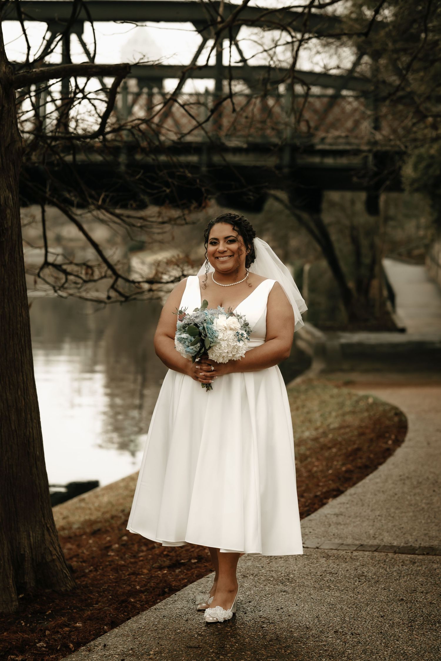 Bride in a flowing white dress poses with bouquet on a gravel path near water.