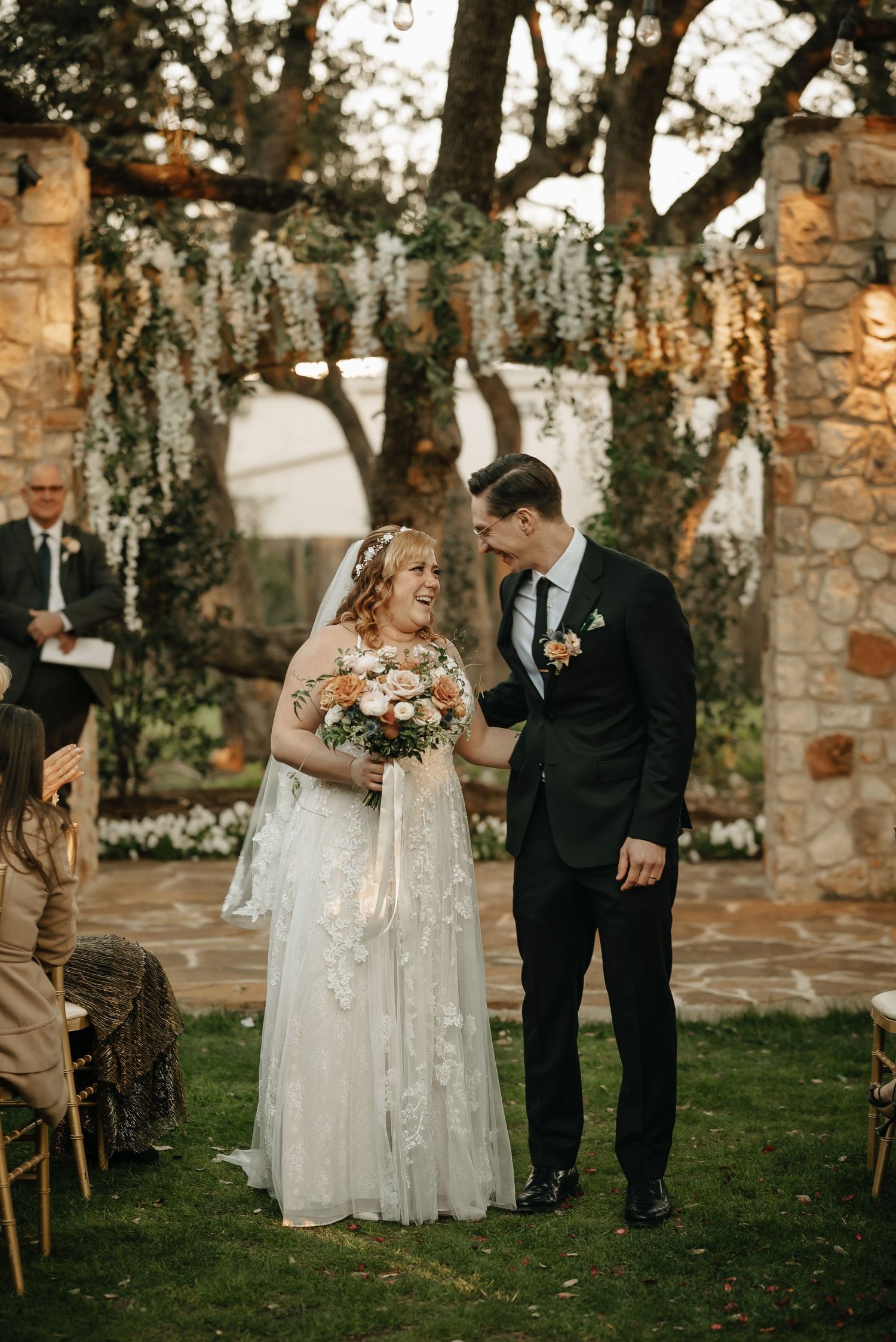 Outdoor wedding ceremony with couple standing under floral arch against rustic stone wall backdrop.