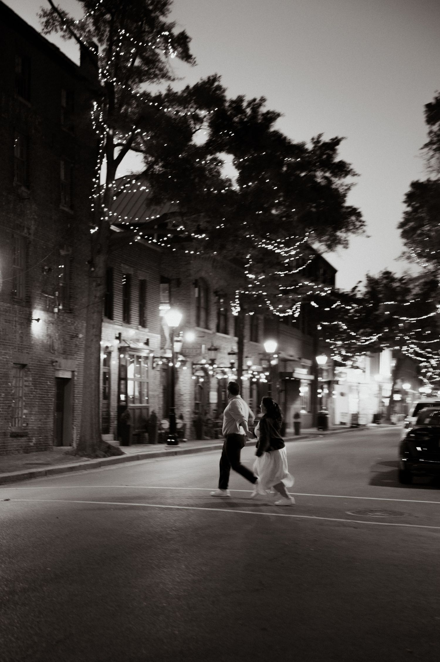 Black and white photo of a couple crossing a street at night, with fairy lights in trees and glowing street lamps.