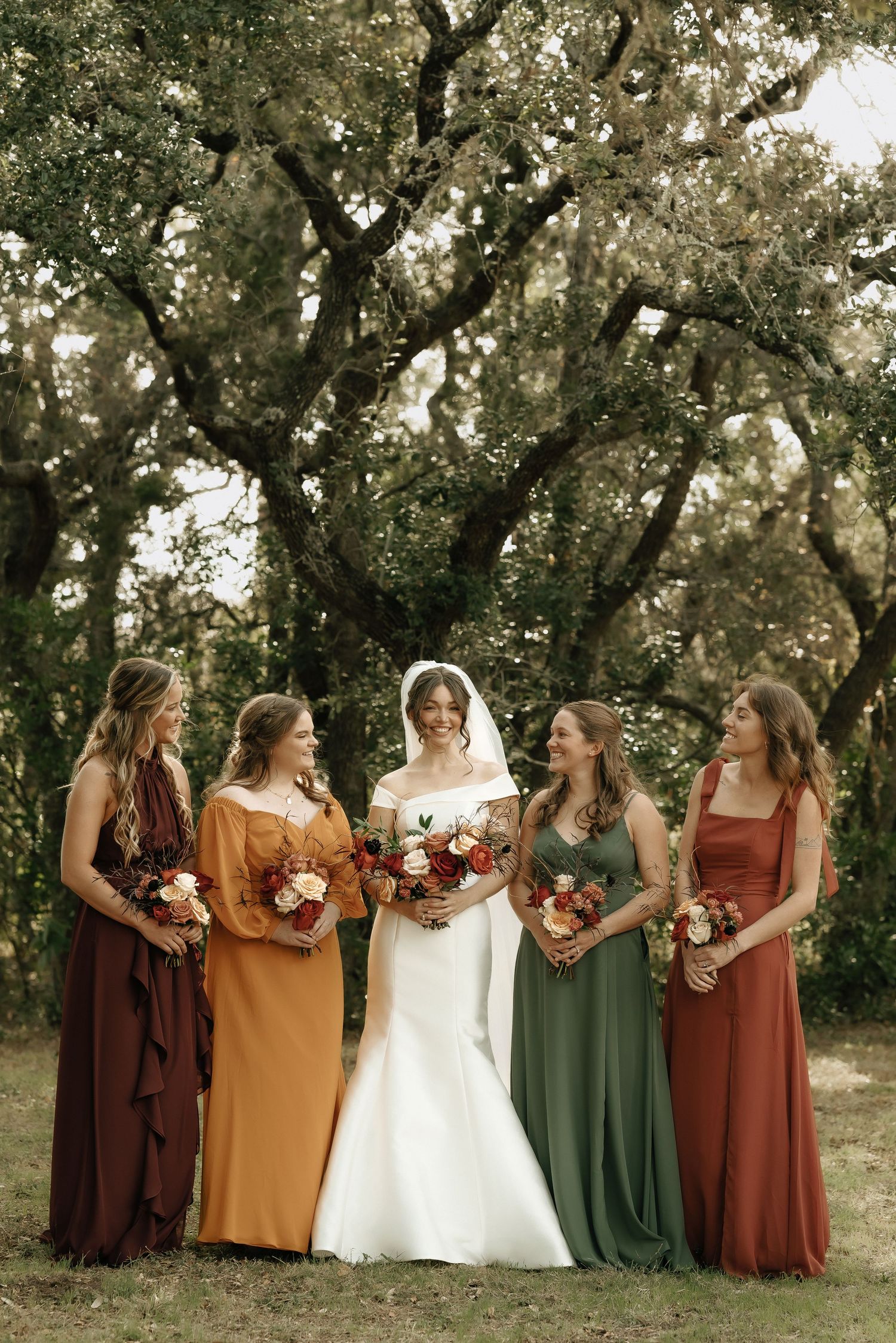 Bridal party poses outdoors with bridesmaids wearing fall-colored dresses against a tree backdrop.