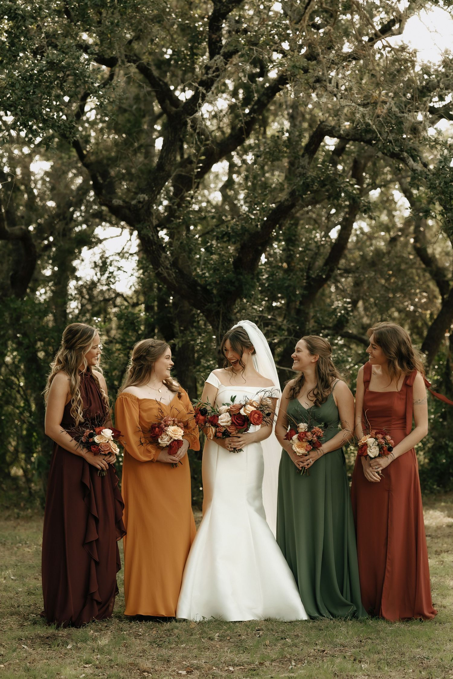 Bridal party in autumn-toned dresses poses together under oak trees.